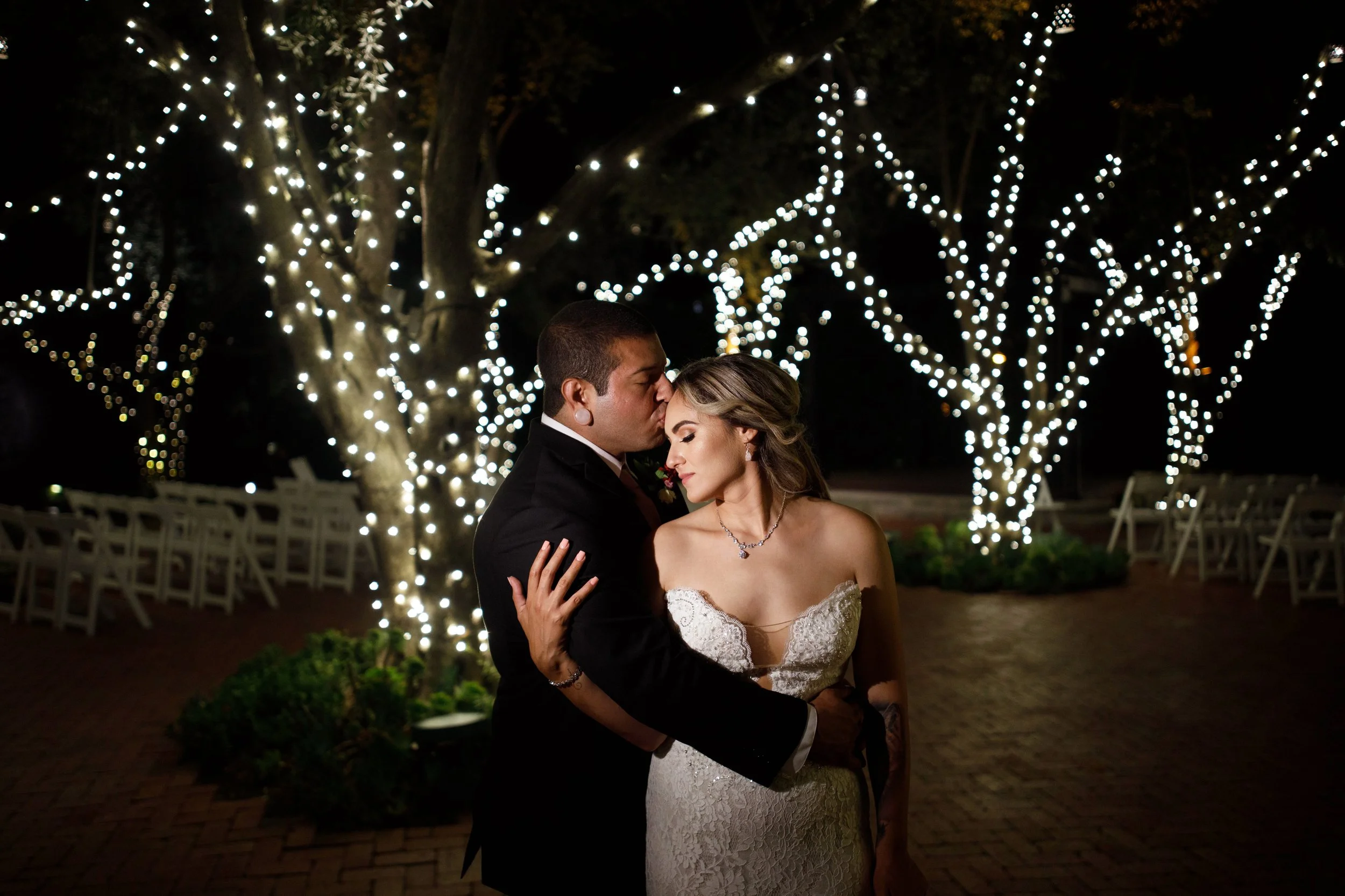A newlywed couple sharing an intimate moment at night, with the groom kissing the bride's forehead. They are outdoors, with trees decorated with string lights in the background creating a romantic ambiance.