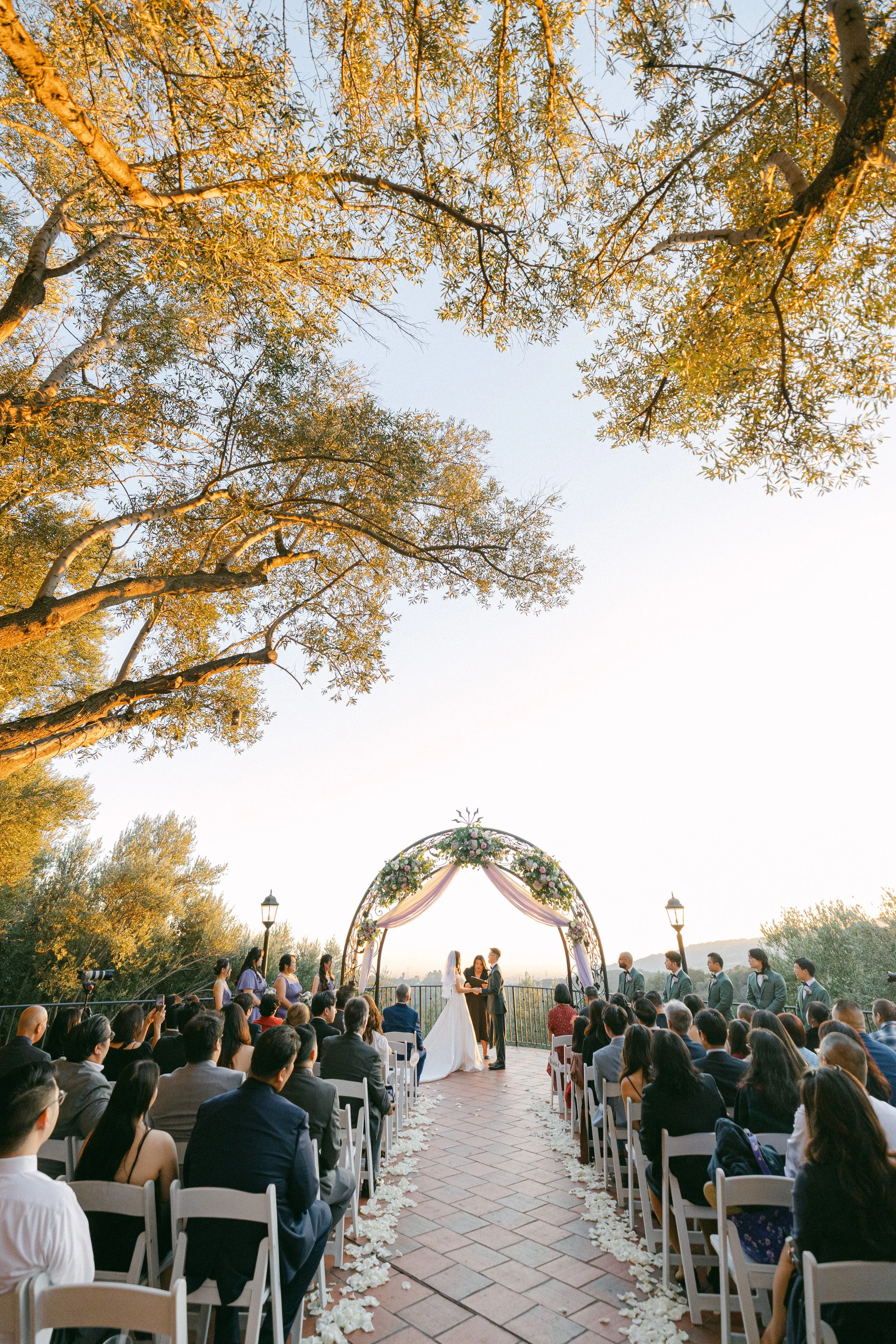 Outdoor wedding ceremony with a bride and groom exchanging vows under a floral arch, surrounded by seated guests, in a natural setting with trees and a clear sky.