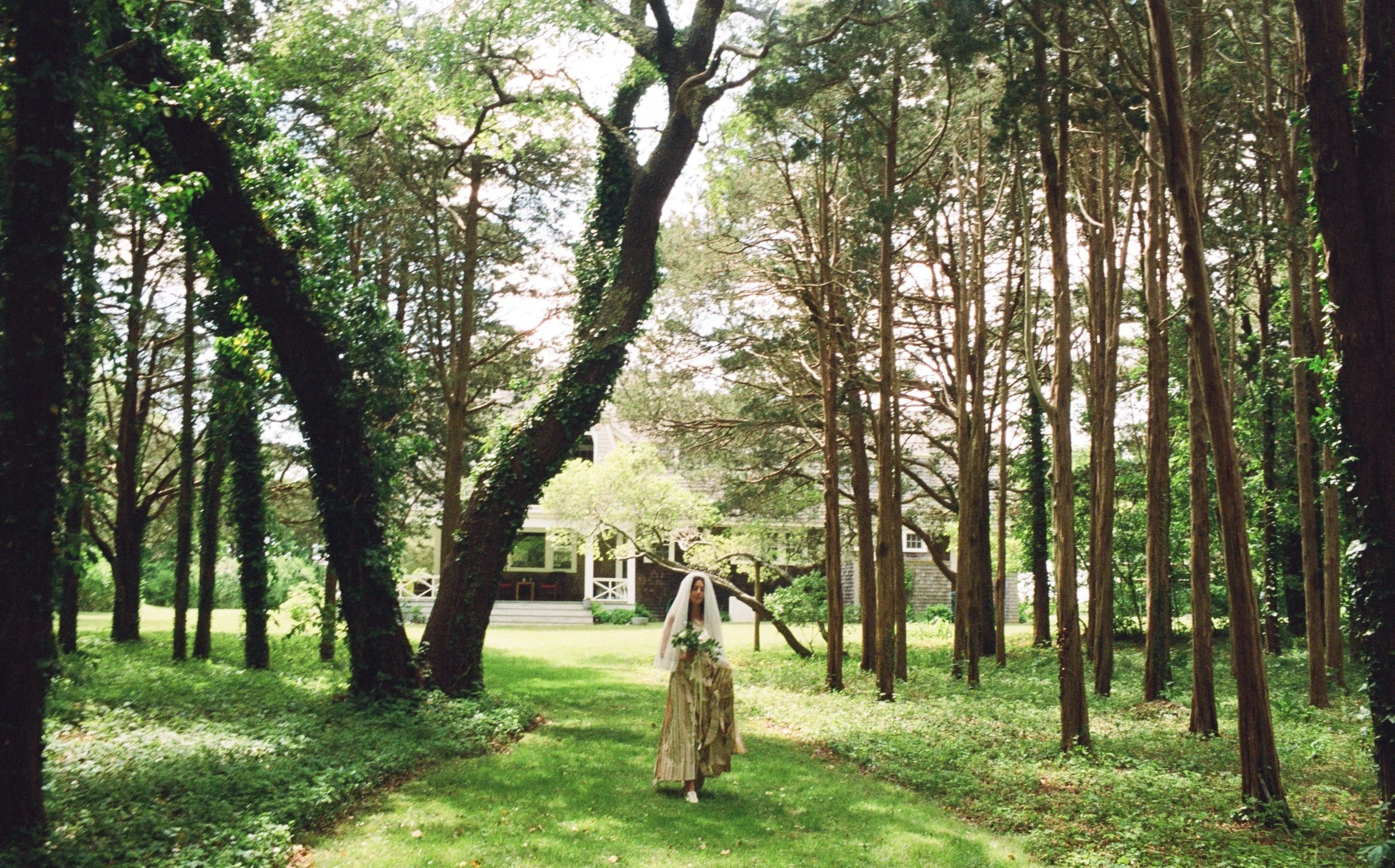 A bride in an artful wedding gown walks down a natural wooded aisle to her outdoor ceremony on Cape Cod Massachusetts.