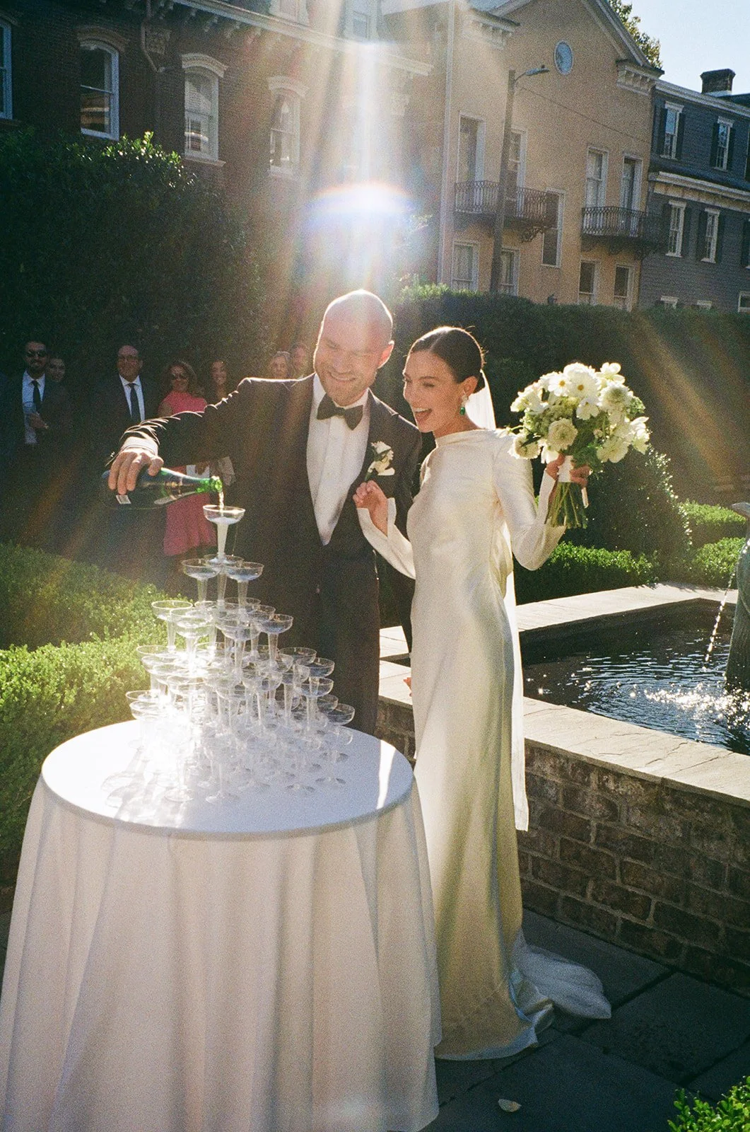 A bride, wearing a silk Danielle Frankel gown, and groom candidly enjoy their champagne tower just after their wedding ceremony in the garden of Harper Fowlkes House in Savannah, Georgia.