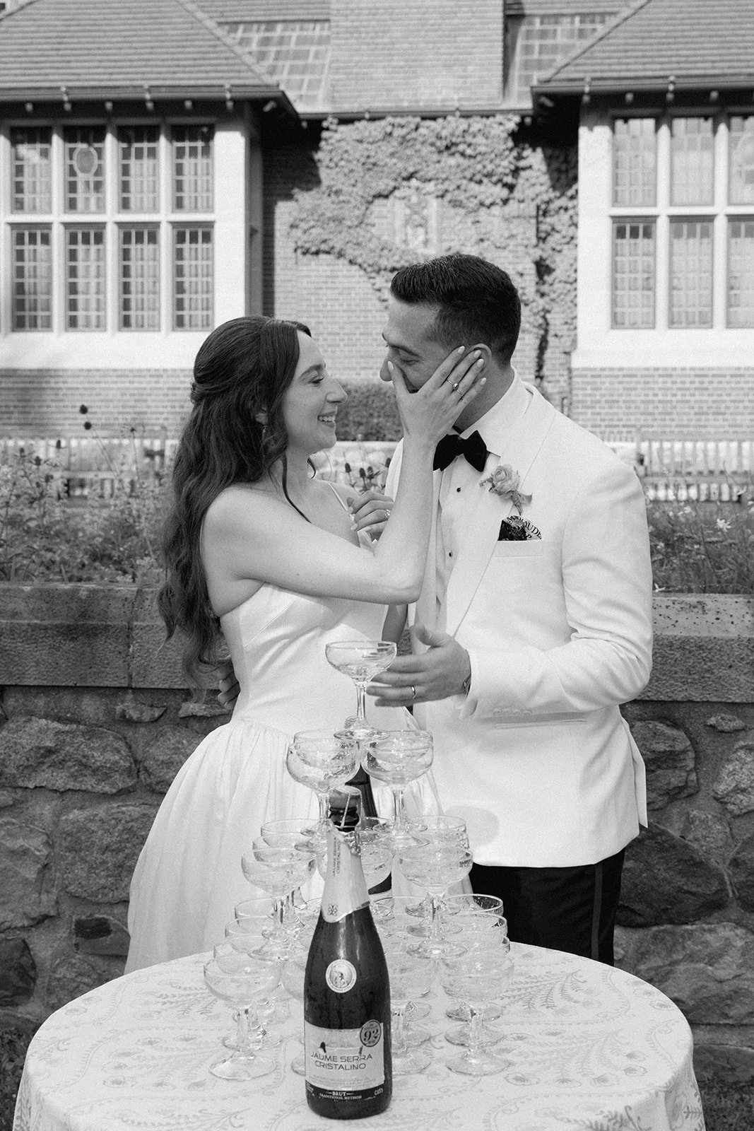Bride wearing Danielle Frankel wipes tear from groom's face during champagne tower pour at Cranbrook House and Gardens outside of Detroit, Michigan.