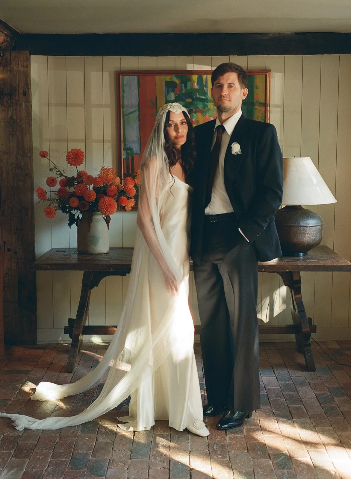 A fashion-forward bride and groom stand for a portrait infront of an organic display of Dahlias in the doorway of their Hudson Valley wedding venue, Stissing House.