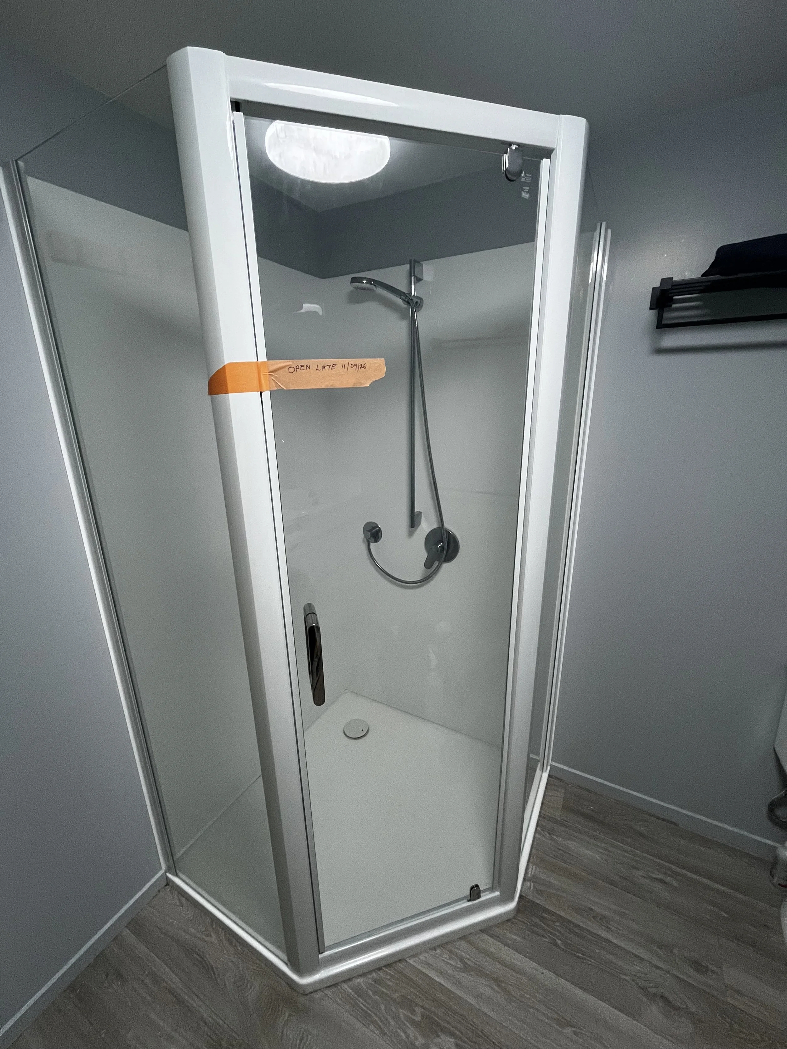 A small enclosed shower stall with a glass door, a handheld showerhead on a vertical rail, a drain on the floor, and a small soap dish holder on the wall, located on a wooden floor next to a gray wall.
