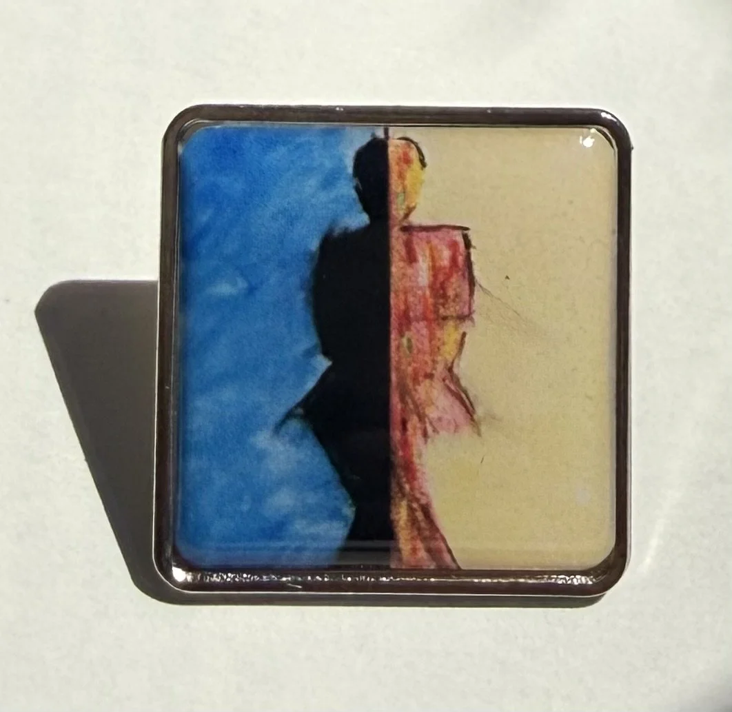 Metal pin with half blue and half yellow background, silhouette of a person in the center, with the blue side showing a shaded figure and the yellow side showing a colorful figure.