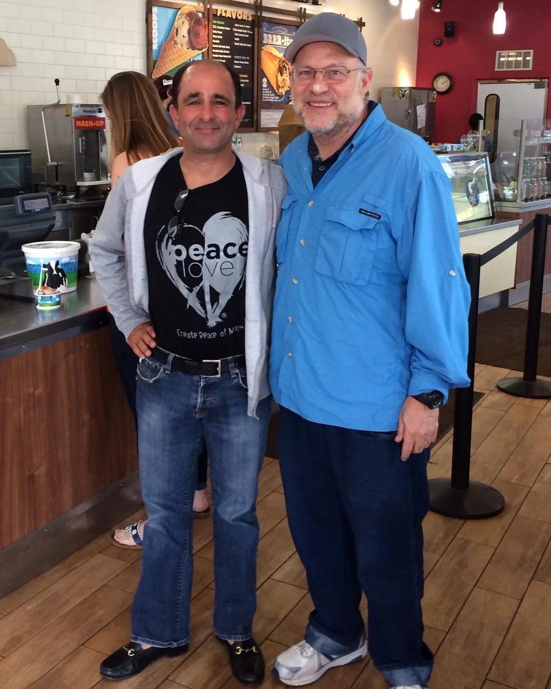 Two men standing inside a cafe, smiling at the camera. One wears a black t-shirt with a peace sign and the words 'peace love', the other wears a blue button-up shirt and a cap. Behind them is a counter with ice cream and a menu board.