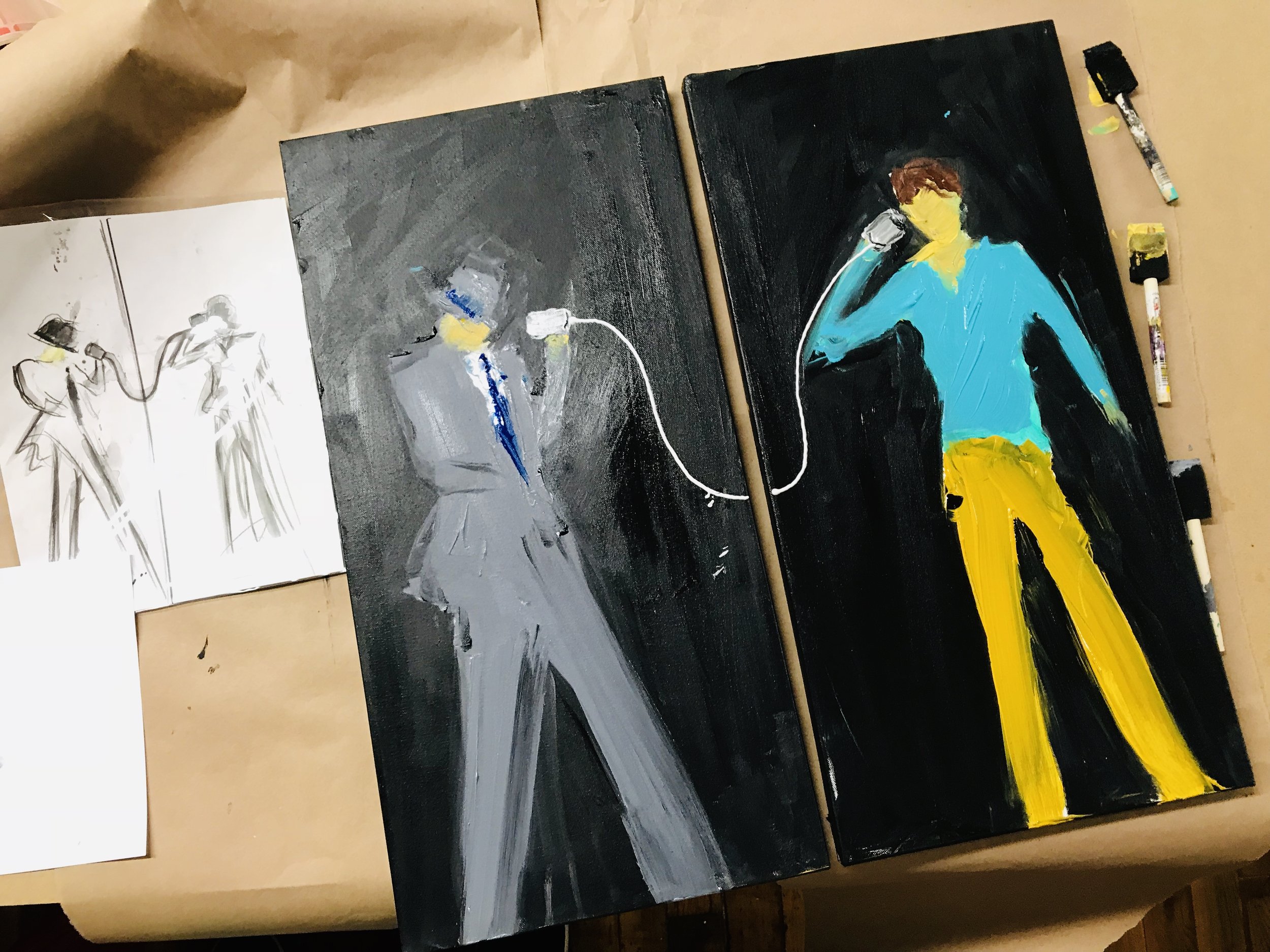 Three abstract paintings on a table, one with a figure in shades of gray and blue, another with a figure in yellow and teal, and the third with bold black and yellow strokes.