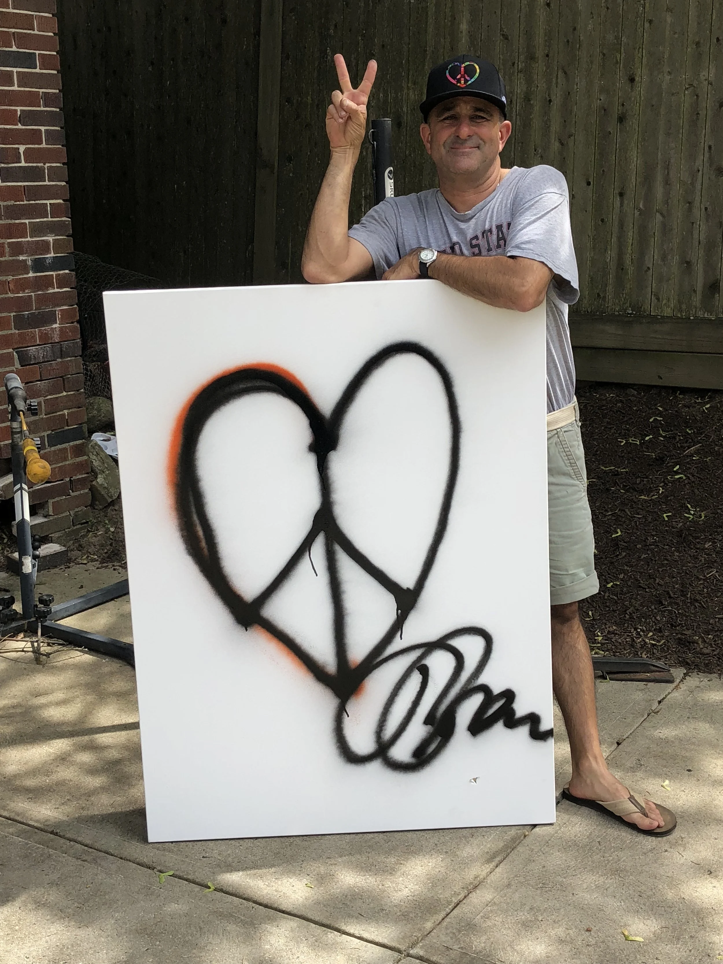 Jeff Sparr wearing a black cap with peace and love symbols, grey T-shirt, khaki shorts, and flip-flops, standing outdoors next to a large white sign with a black graffiti heart and word, showing a peace sign with his hand.