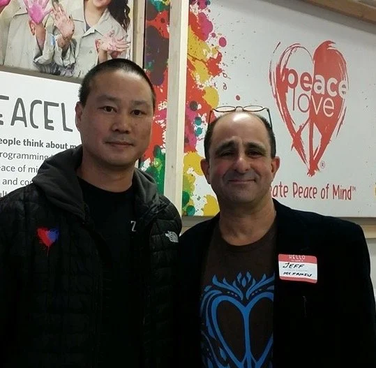 Two men standing side by side smiling in front of a Peace Love sign and colorful background at an event.