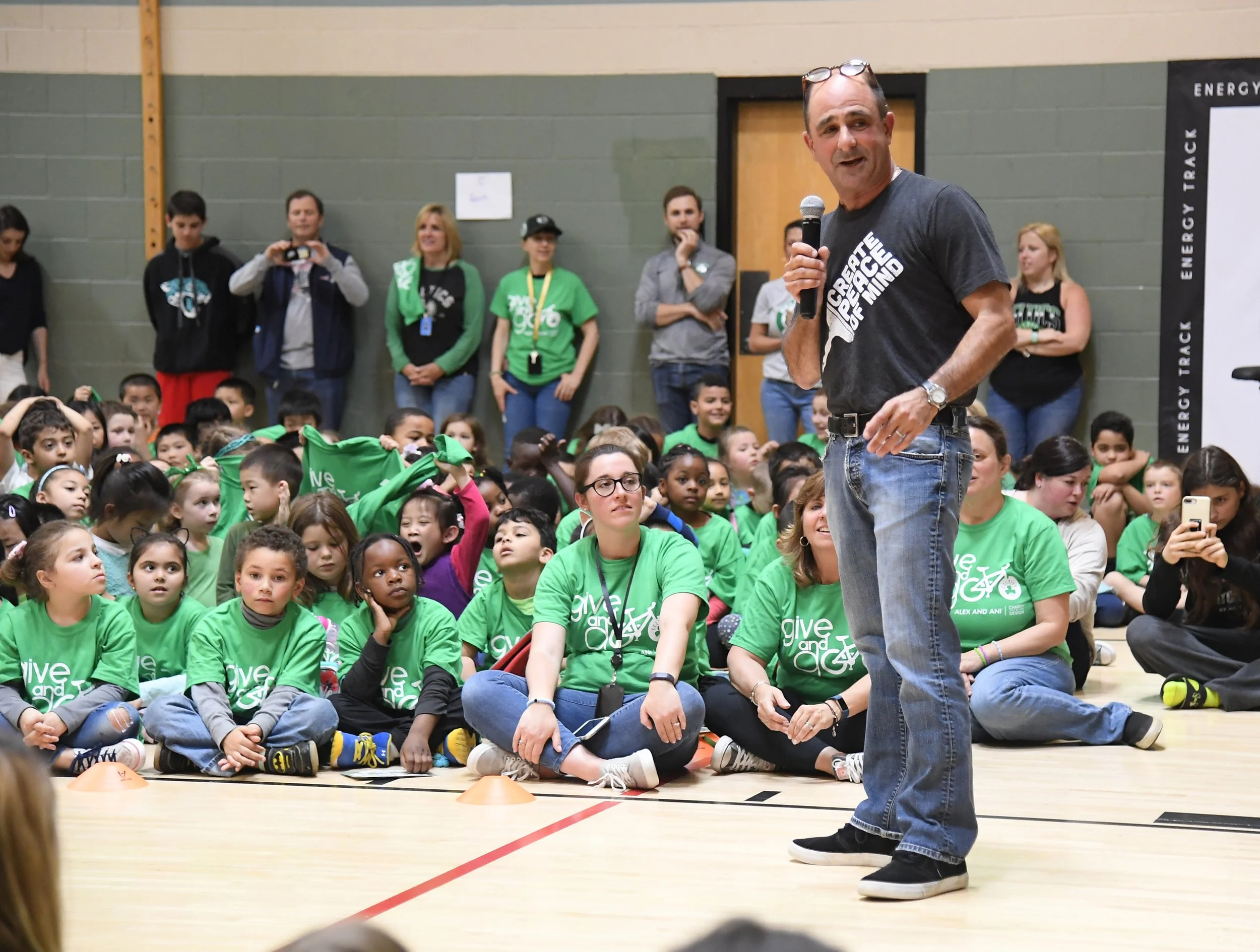 Jeff Sparr holding a microphone speaking to a group of children sitting on the floor in a gymnasium, with several adults standing behind them, all wearing green shirts with the words "give and go."