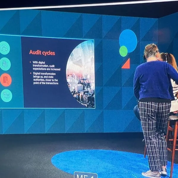 People attending a presentation or conference in front of a large screen displaying a slide about audit cycles and digital transformation.