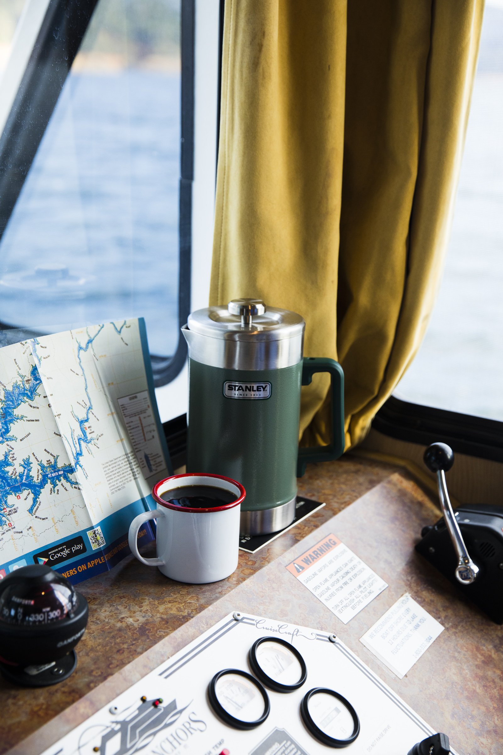 A table inside a boat with a cup of coffee, a green Stanley thermos, a map, and some documents near a window with yellow curtains.