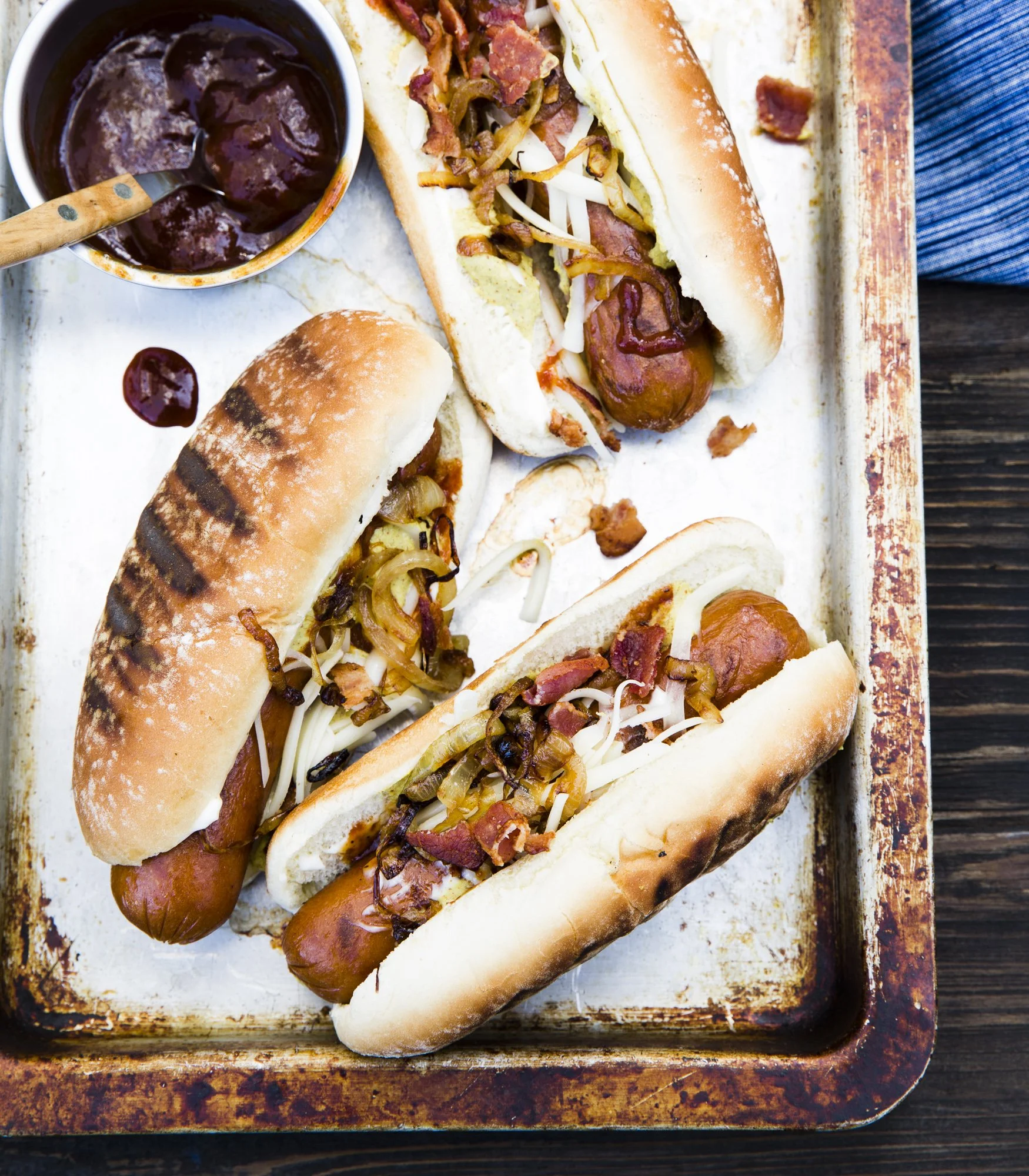 Three hot dogs in buns topped with grilled onions and bacon, served on a baking sheet, with a side of barbecue sauce.