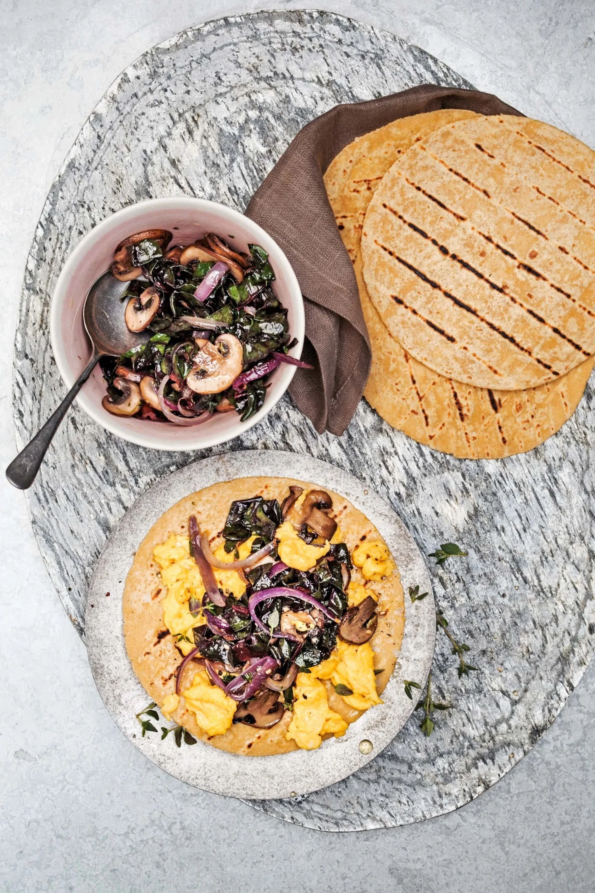 A plate with scrambled eggs, mushrooms, and onions on a tortilla, a bowl of sautéed mushrooms and spinach with onions, and three grilled tortillas on a napkin on a rustic wooden table.
