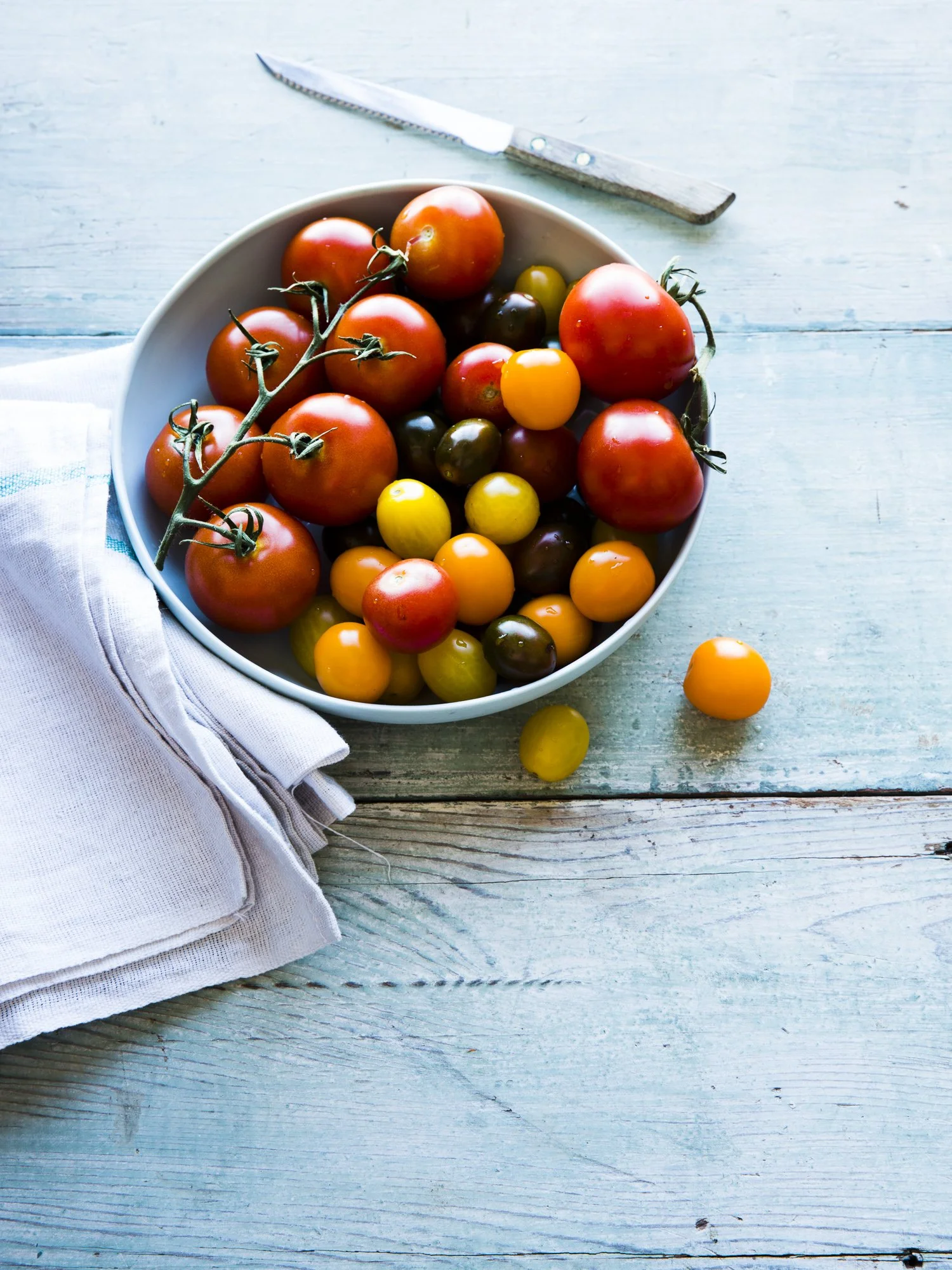 A bowl of assorted heirloom tomatoes on a light blue wooden surface, with a knife and a white cloth nearby.