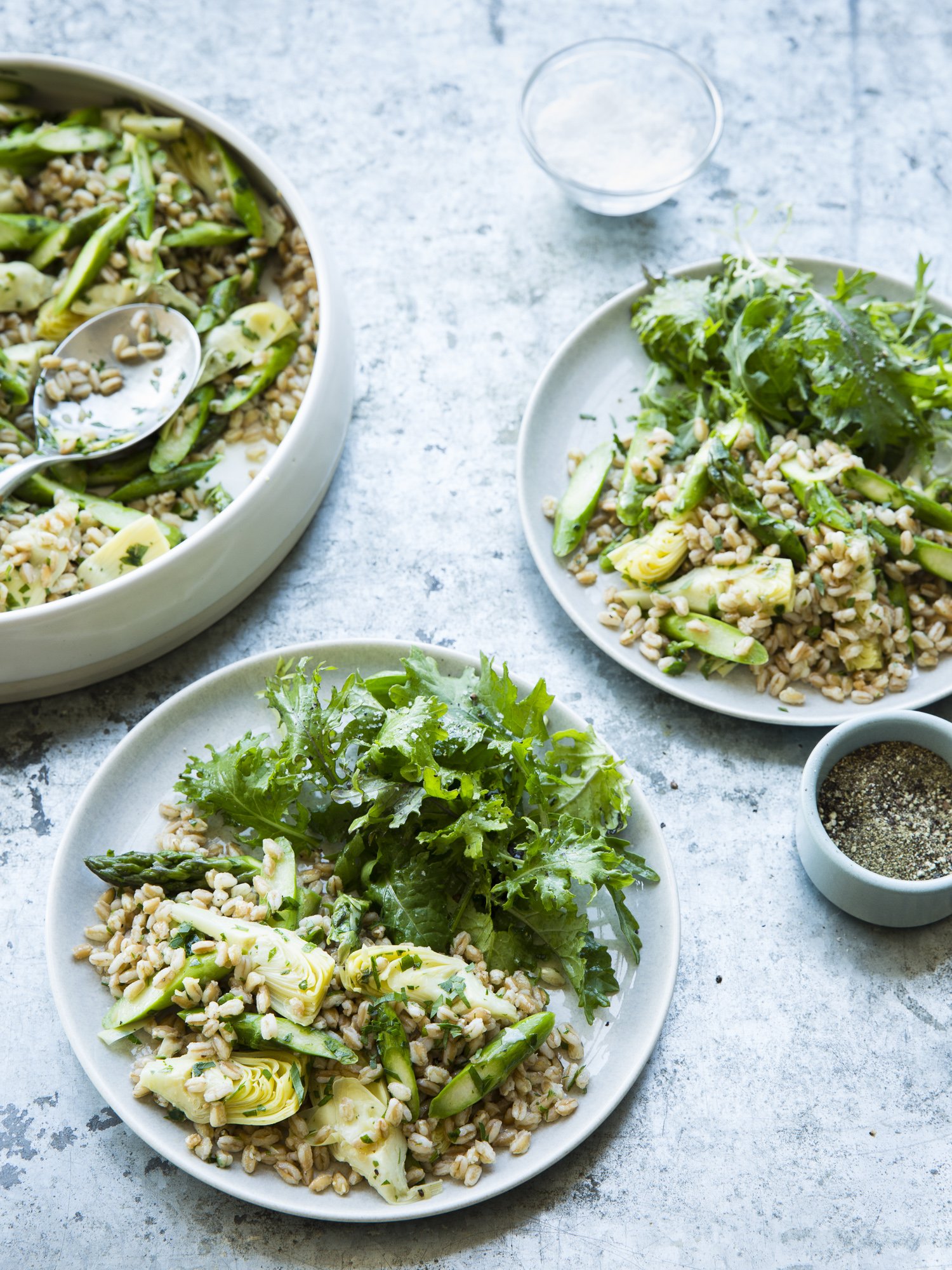 Salad bowls with grains, sliced zucchini, artichokes, and mixed greens on a light textured surface.