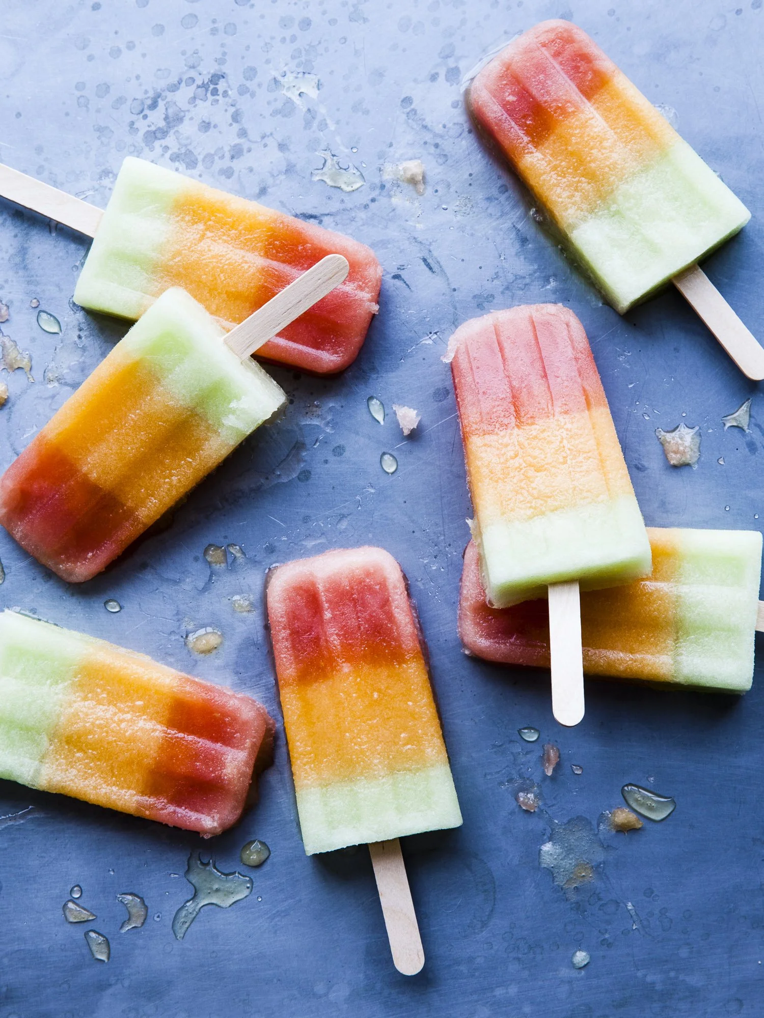 Colorful watermelon popsicles on a blue surface, some with popsicle sticks, with melting ice and water around them.