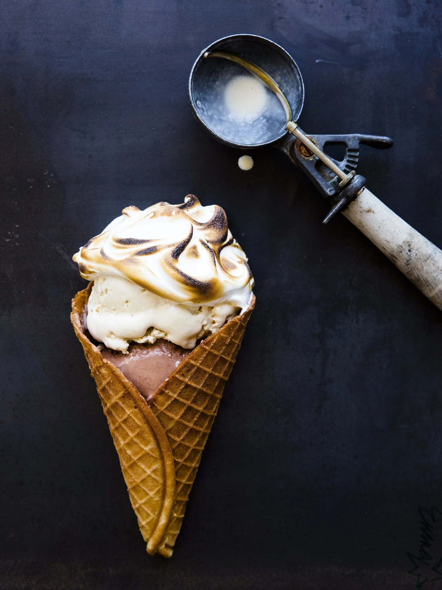 Scoops of ice cream in a waffle cone with a marshmallow topping, and a vintage ice cream scoop resting on a dark surface.
