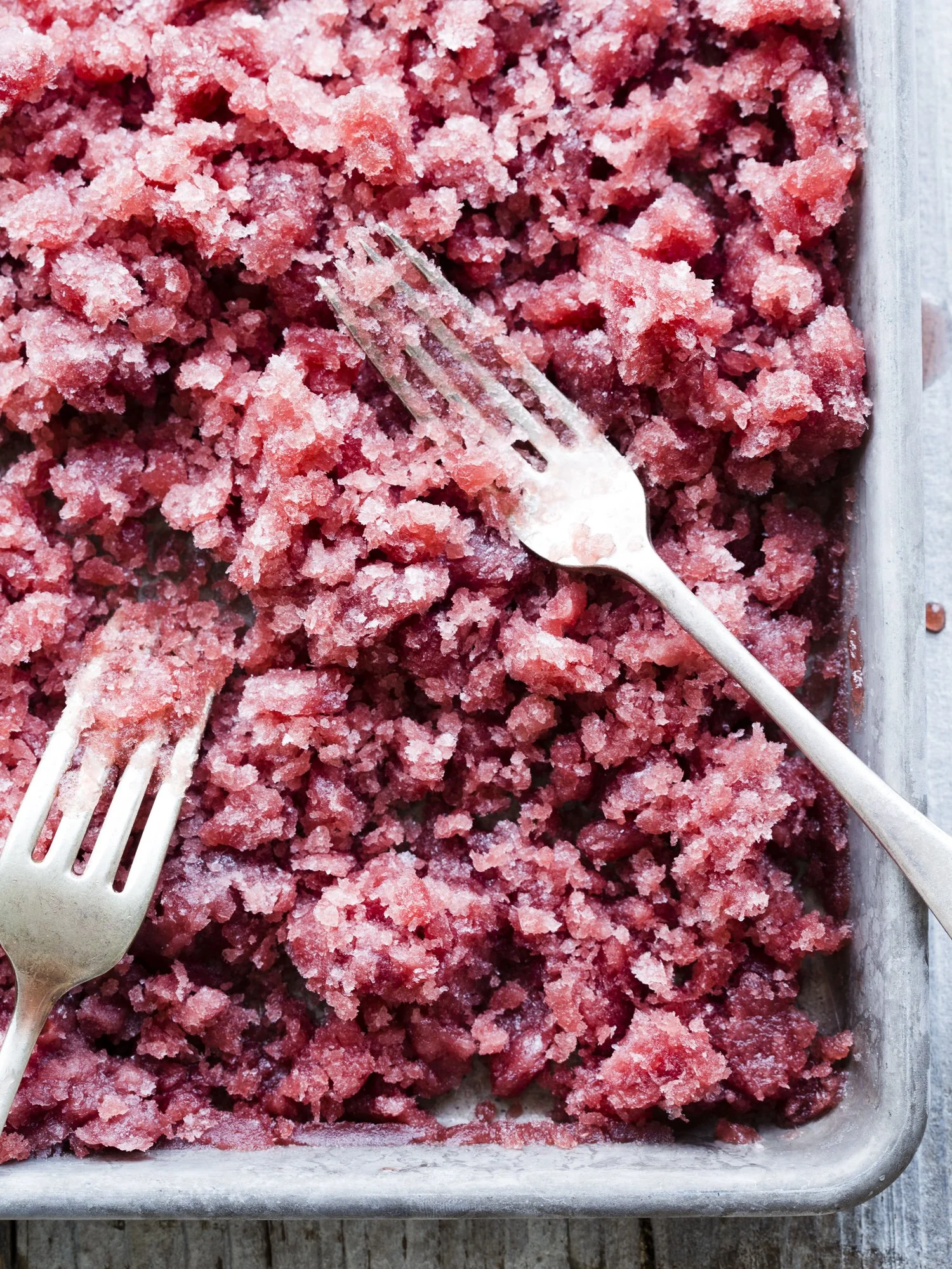 Close-up of a metal tray filled with frozen pink crushed berries, with two metal forks resting on top.