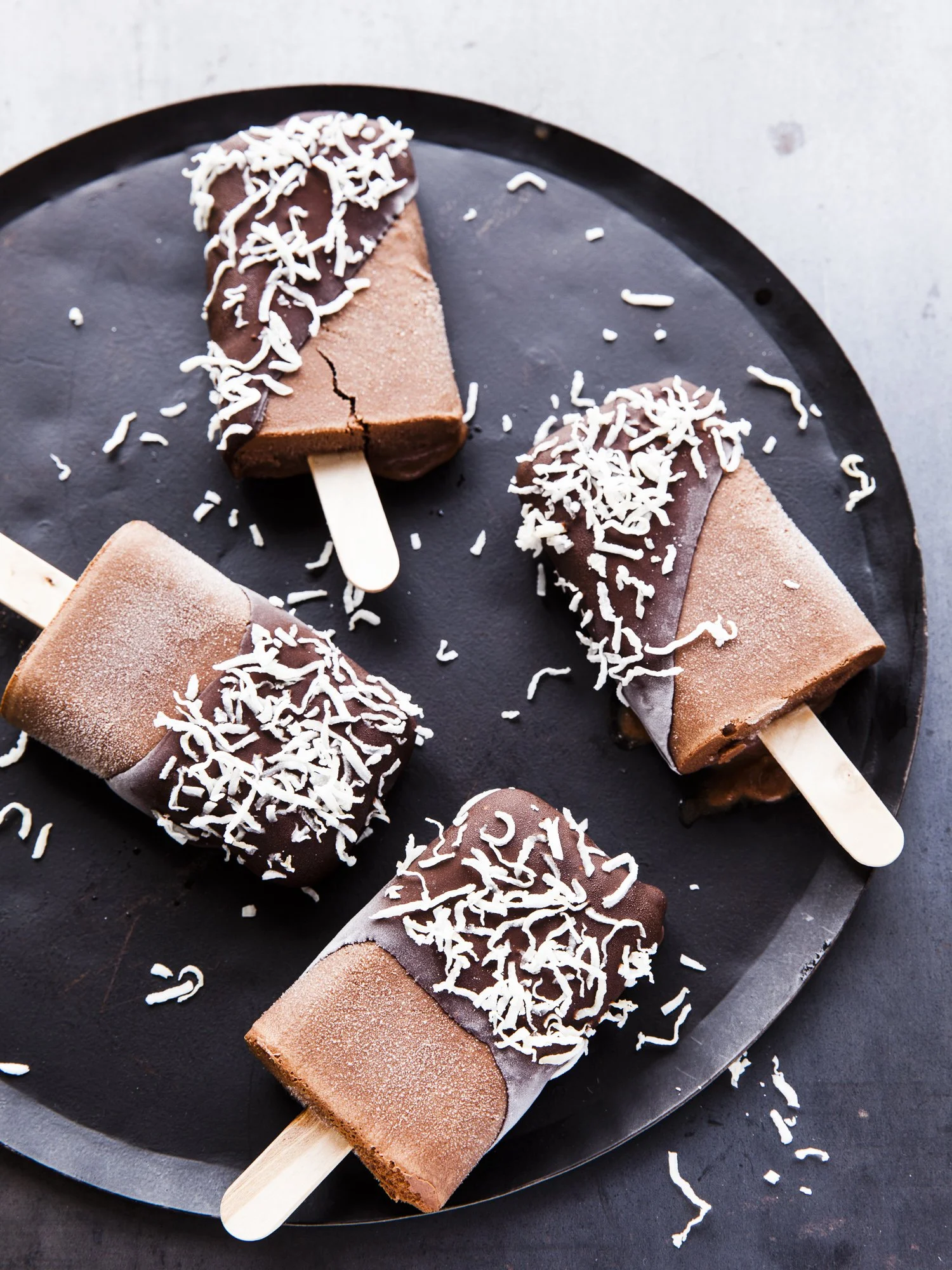 Four chocolate popsicles with shredded coconut on a black round plate.