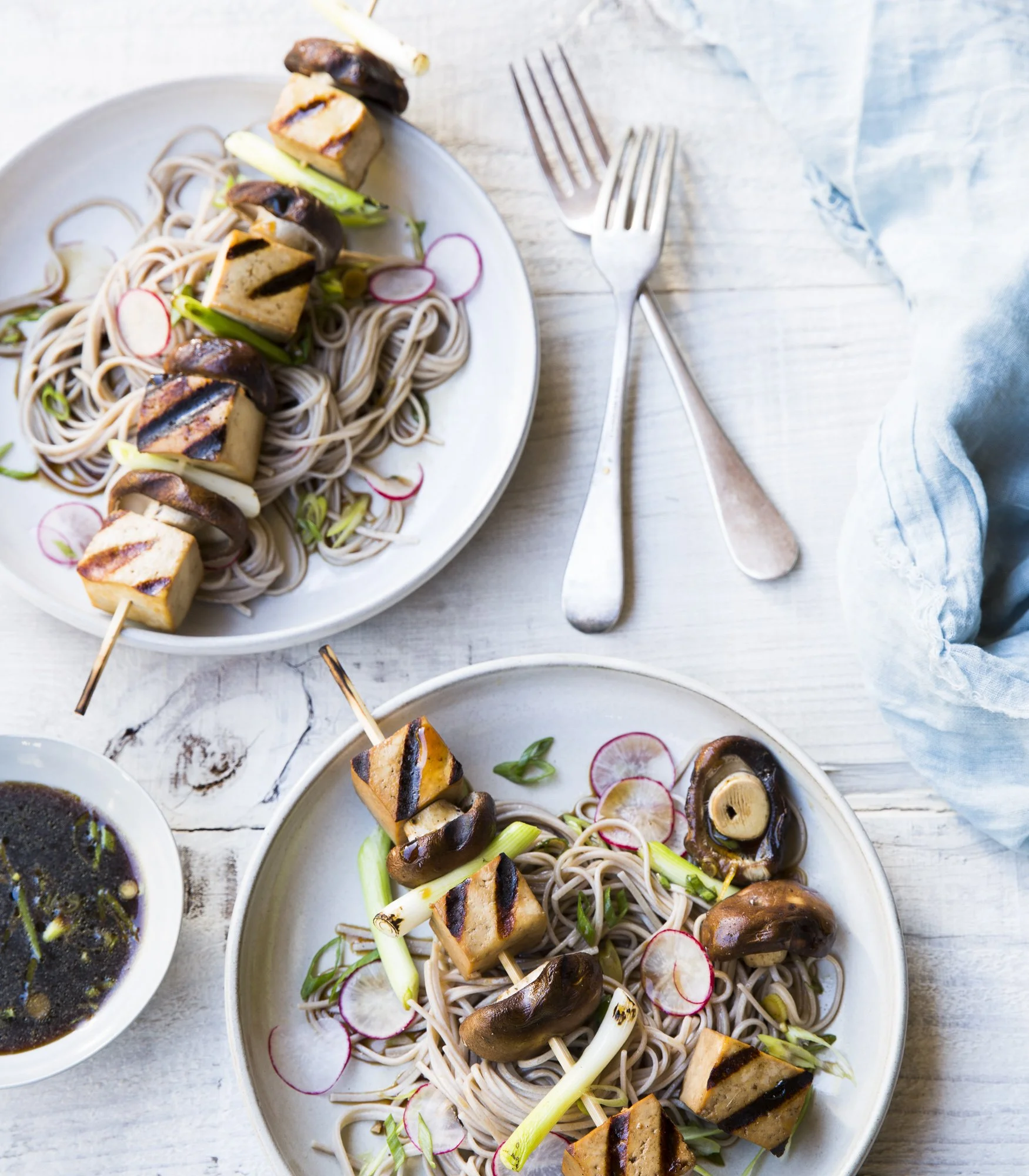 Two bowls of soba noodle salads with grilled tofu, mushrooms, sliced radishes, and green onions on a white wooden table, with forks and a small bowl of dipping sauce.