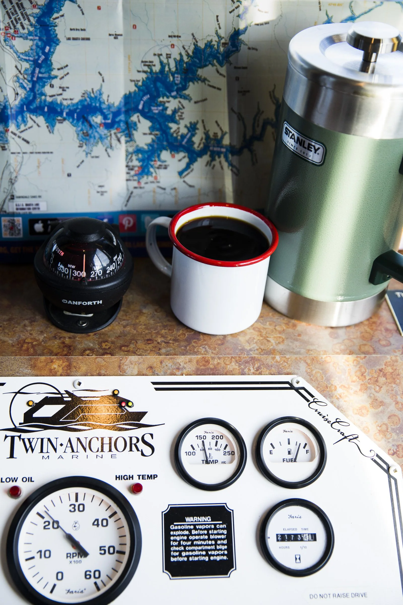 A map in the background, a white mug filled with black coffee, a green Stanley thermos, a small black Danforth compass, and a Twin Anchors Marine control panel with gauges and meters on a wooden surface.
