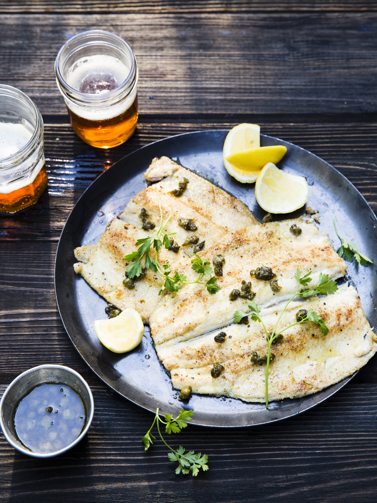 Grilled fish fillet garnished with fresh parsley and capers, served with lemon wedges, accompanied by two glasses of beer, on a dark wooden table.