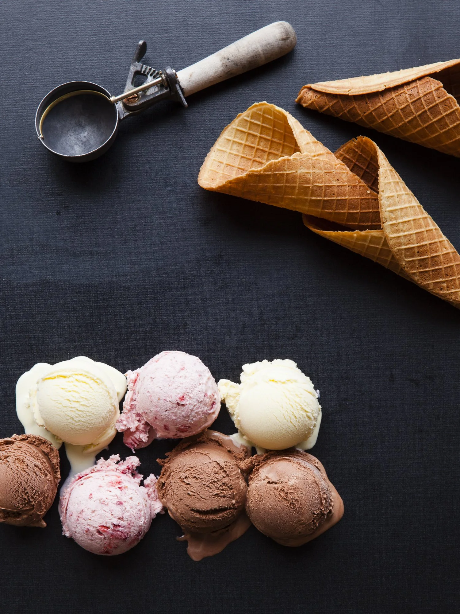 Multiple ice cream scoops in various flavors placed on a dark surface with waffle cones and an ice cream scooper nearby.