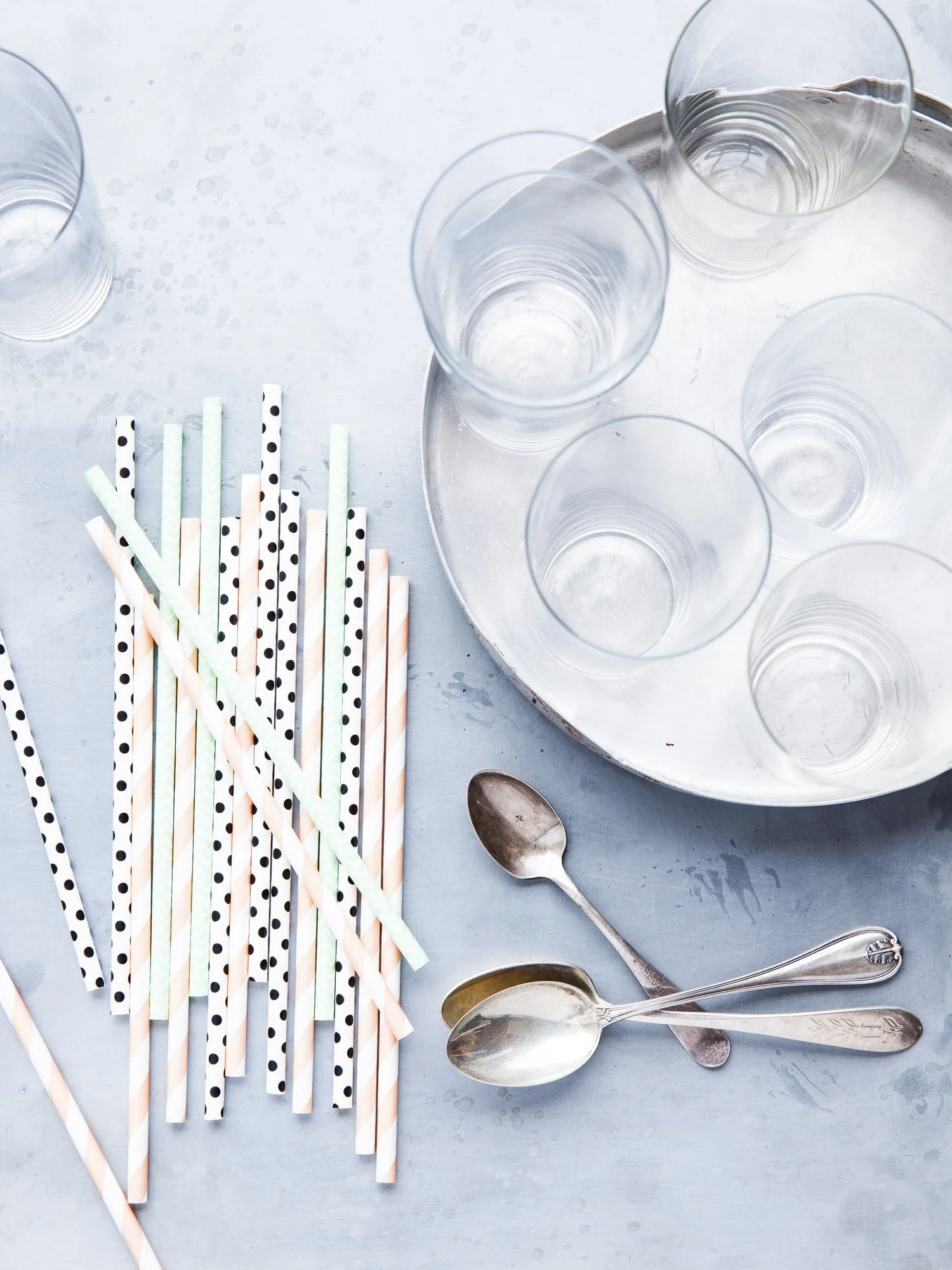Clear glasses on a silver tray, colorful striped and polka dot paper straws, and vintage spoons on a light blue surface.