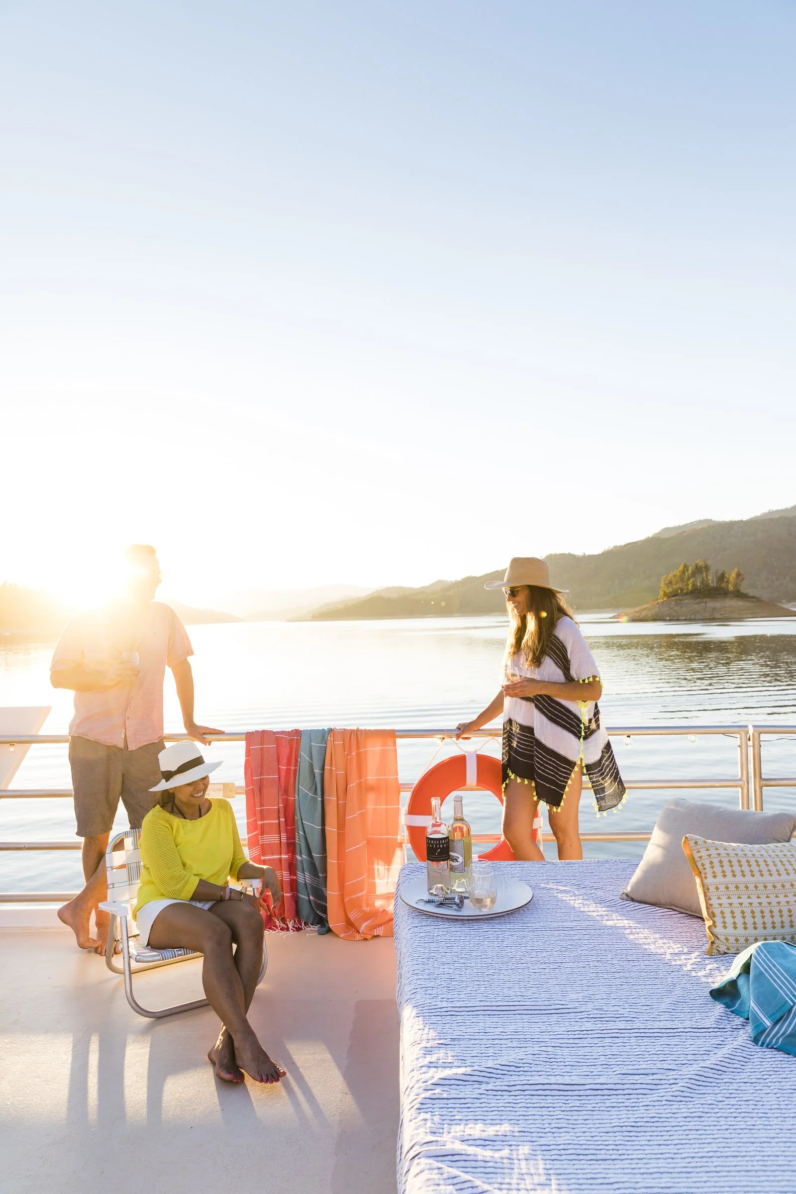 Three women and a man enjoying a sunset on a boat, with drinks on a table, wearing summer clothes and hats, near a body of water and mountains in the background.