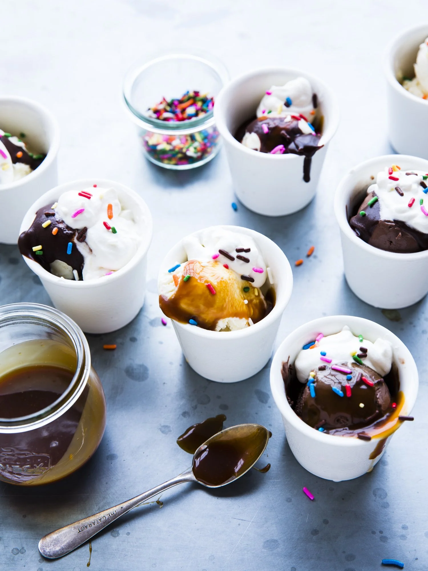 Multiple cups and jars filled with ice cream, chocolate, caramel sauce, and colorful sprinkles on a metallic surface.
