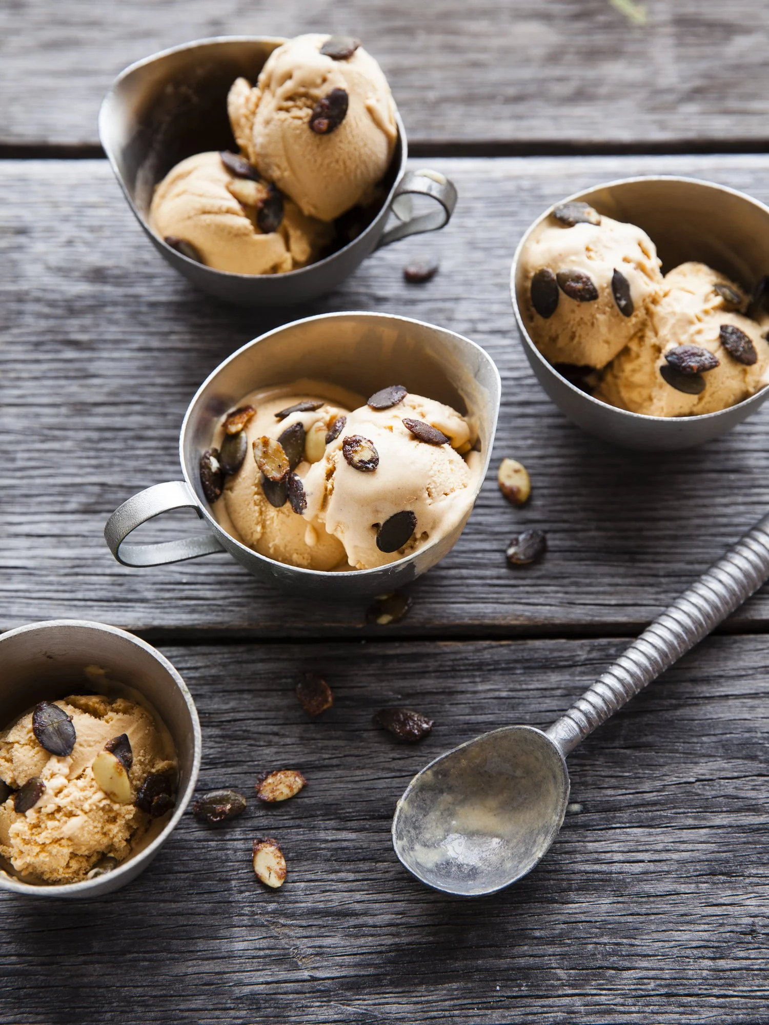 Four small metal cups filled with vanilla ice cream topped with chocolate chips, placed on a rustic wooden surface with scattered chocolate chips and a vintage metal ice cream scoop nearby.