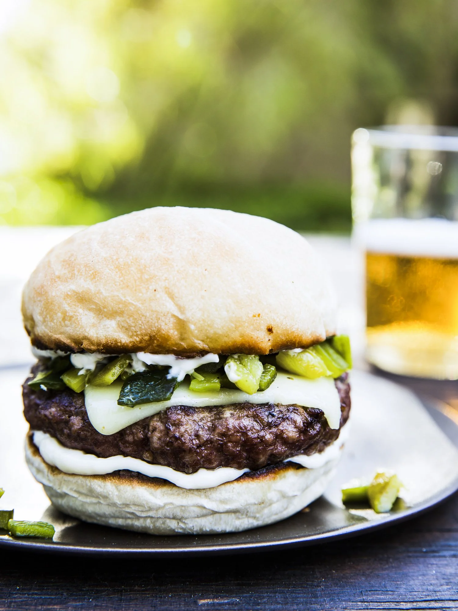 Close-up of a hamburger with beef patty, cheese, pickles, and sauce in a bun, with a glass of beer in the background.