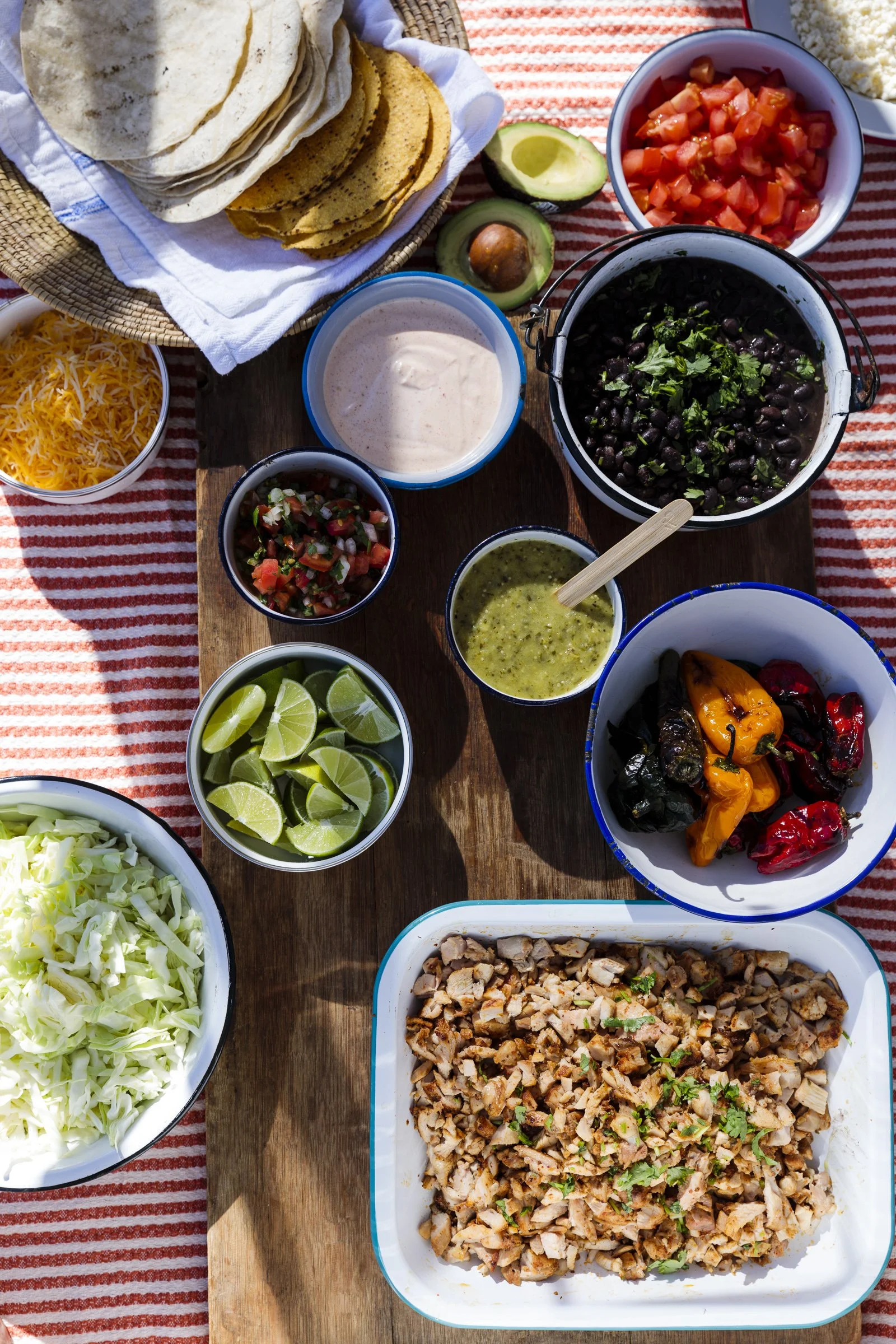 A colorful Mexican-style food spread on a wooden table, including chopped grilled chicken, shredded cheese, chopped tomatoes, chopped lettuce, lime wedges, sliced avocados, various salsas, black beans, grilled peppers, a creamy sauce, and tortillas i