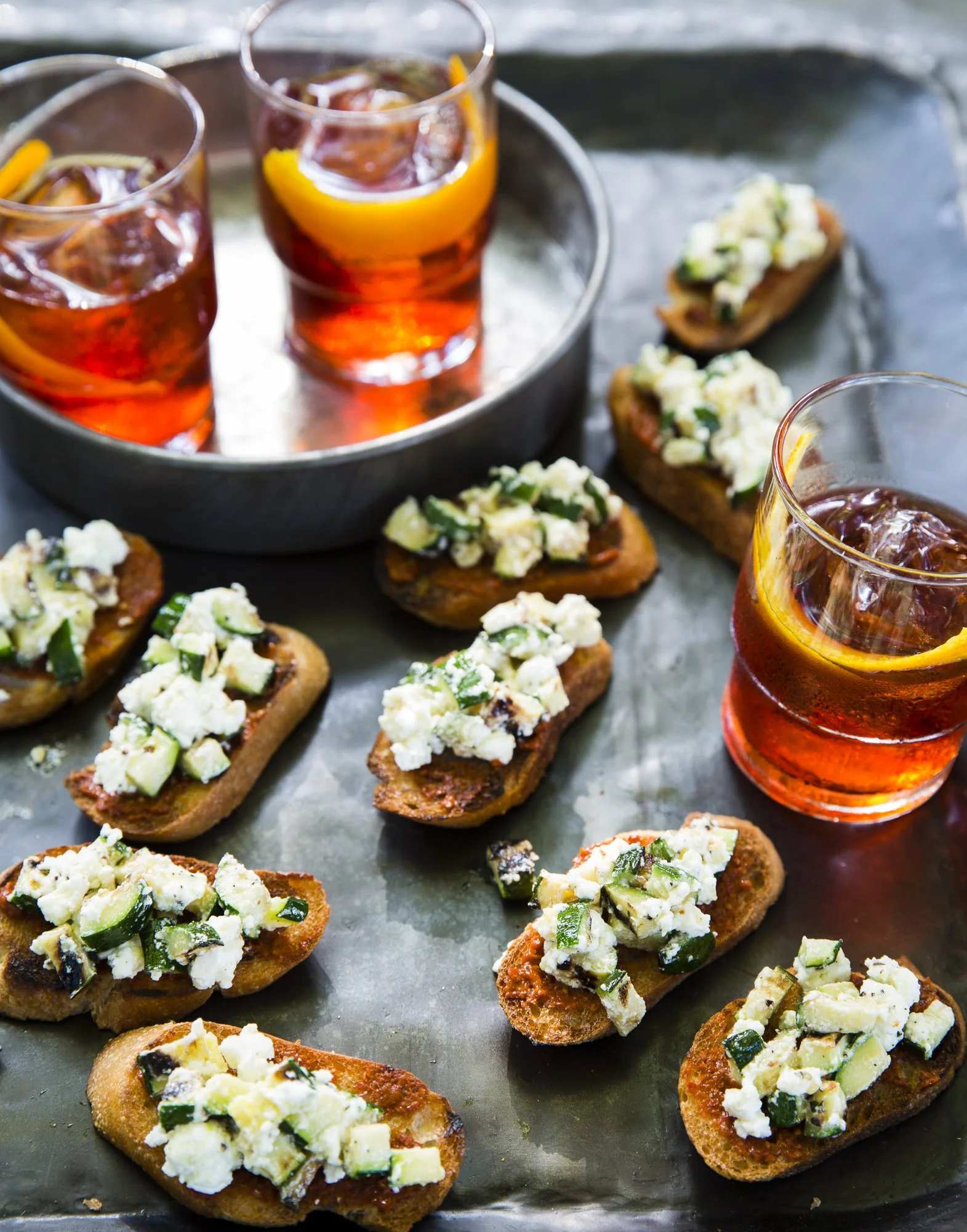 Close-up of toasted bread slices topped with crumbled blue cheese and chopped cucumbers, served with glasses of iced tea with lemon slices.