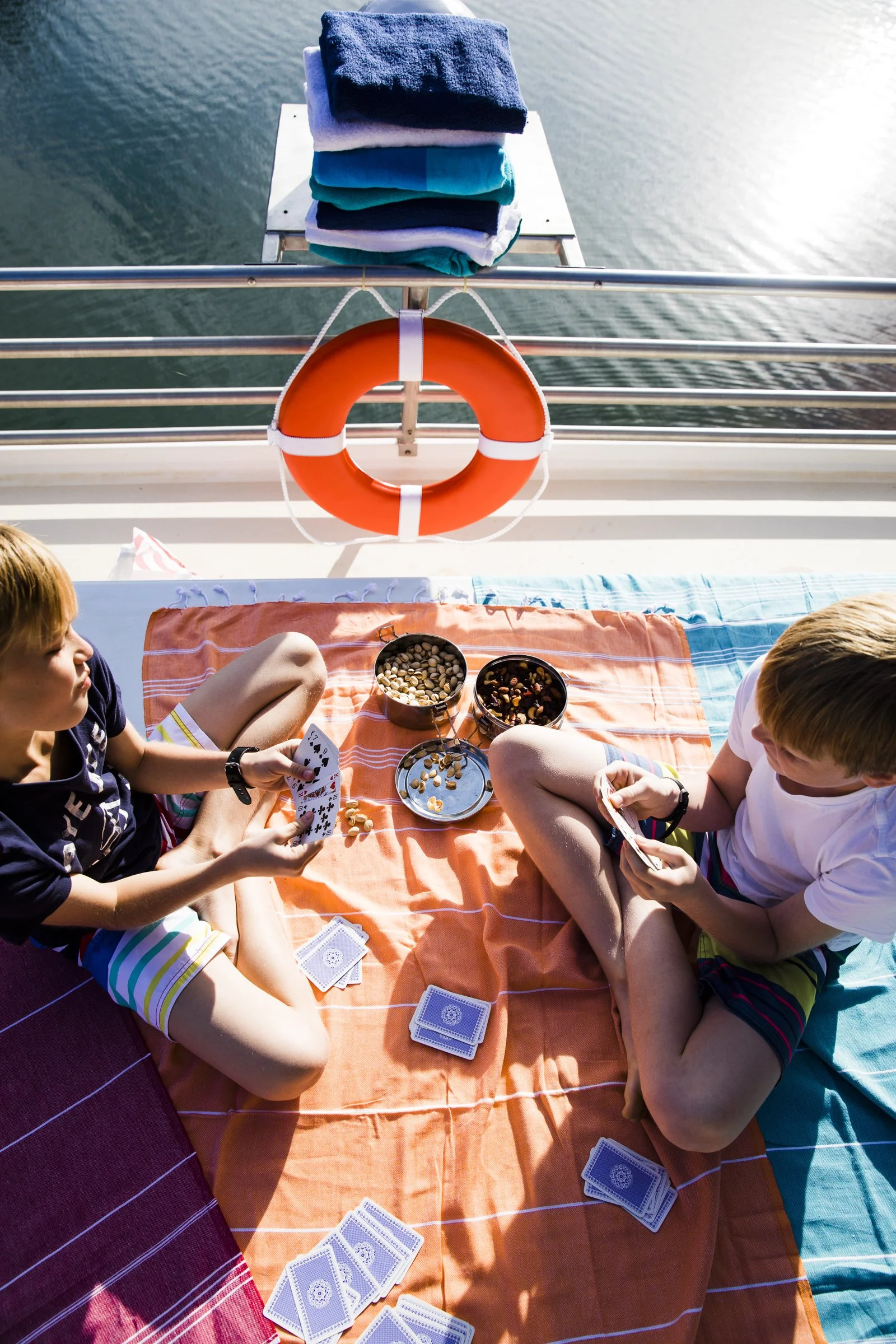 Two boys playing cards on deck of boat with water in background, towels, life preserver, and folded clothes on railing.