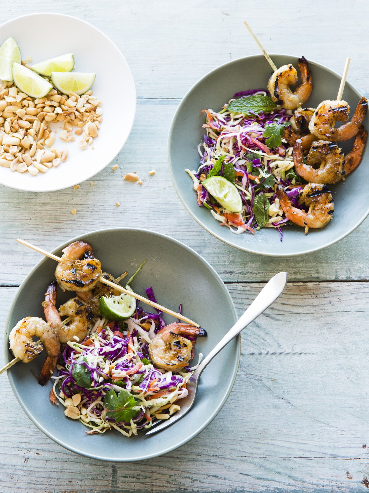 Two bowls of grilled shrimp with coleslaw and lime wedges, with a plate of chopped peanuts and lime wedges nearby on a light wooden table.