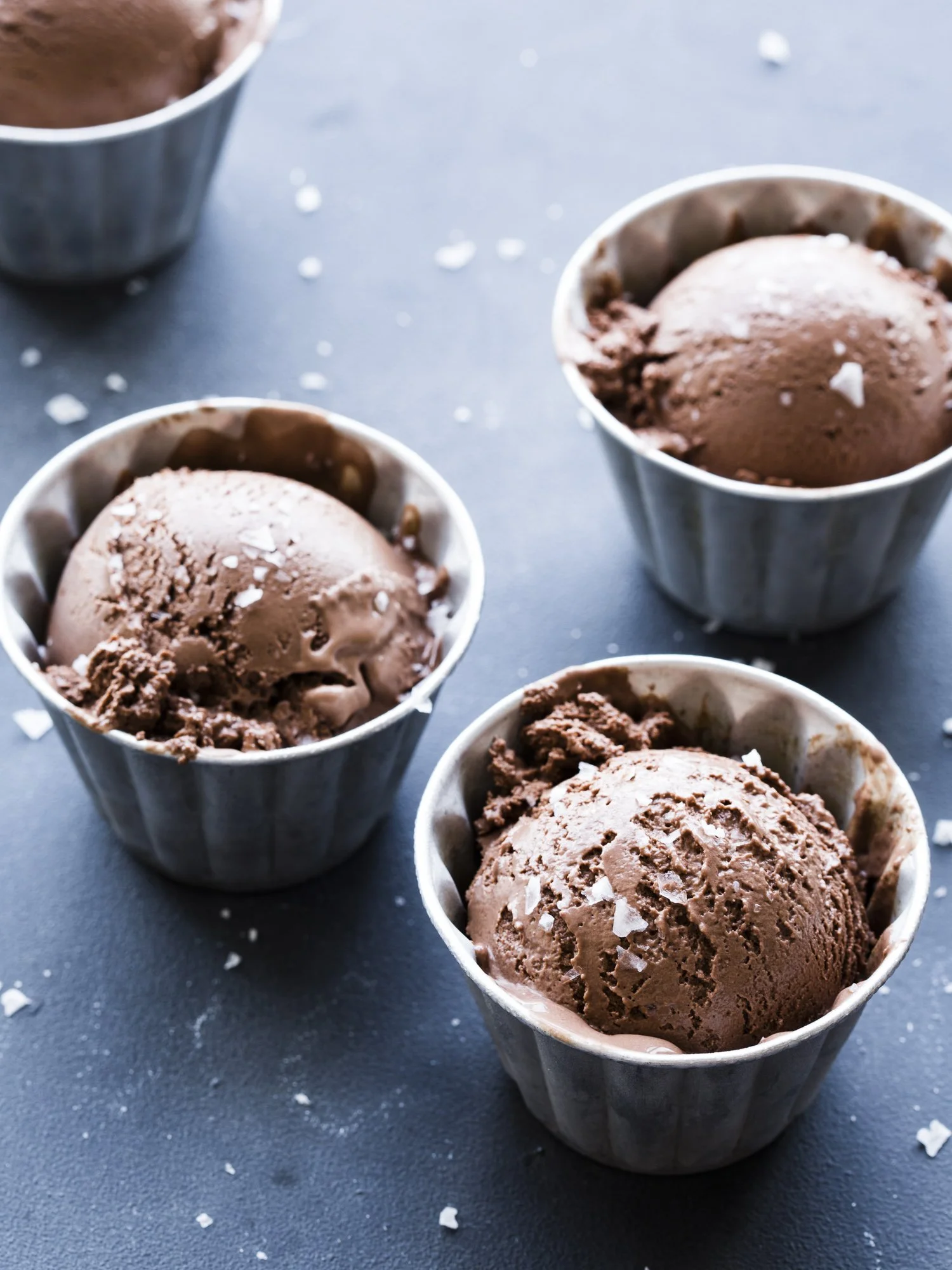Four cups of chocolate ice cream with sprinkled salt flakes on top, placed on a dark surface with scattered salt flakes.