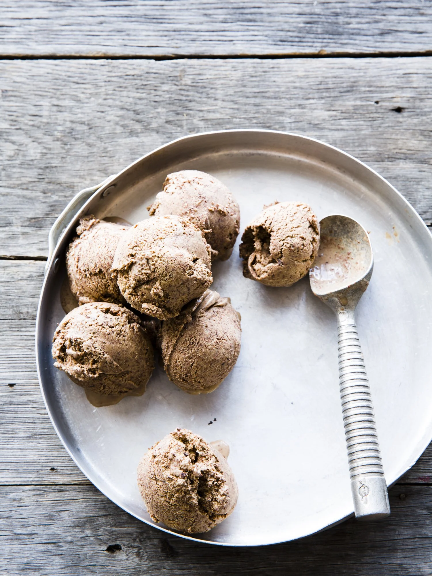 Scoops of chocolate ice cream on a metal tray with a metal ice cream scoop beside them, on a wooden surface.
