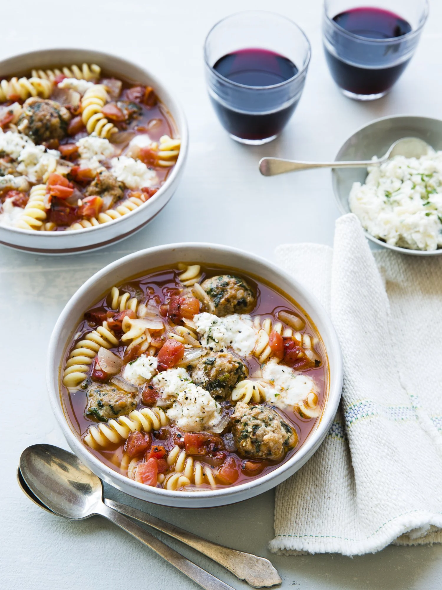 Bowls of Italian soup with pasta, meatballs, tomatoes, and cheese, accompanied by glasses of red wine and a side of mashed potatoes.
