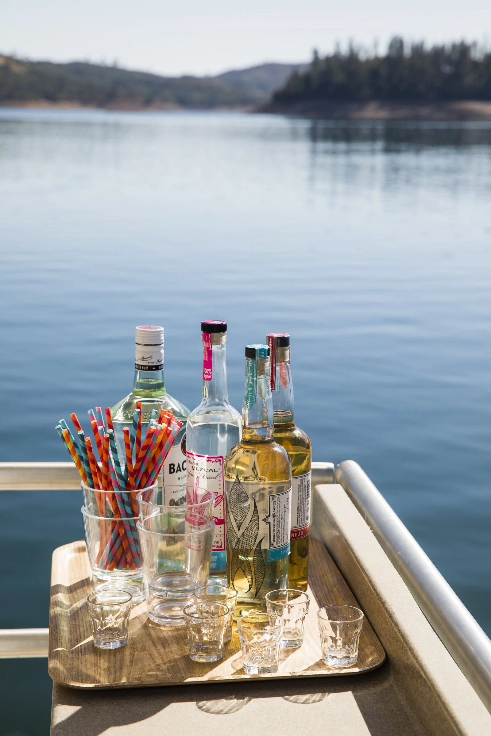 A tray with bottles of alcohol, shot glasses, and colorful straws on a boat deck overlooking a lake with hills in the background.