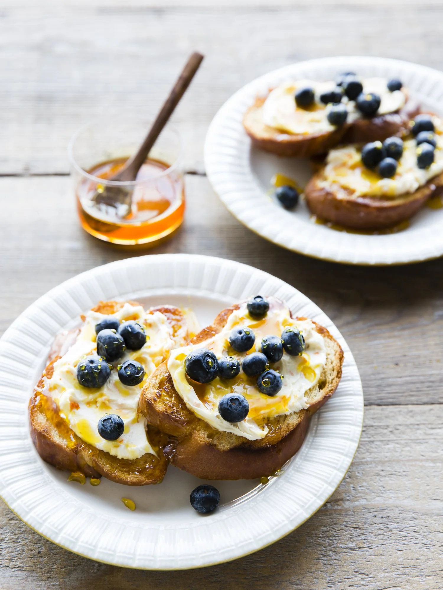 Two plates with bread topped with eggs, blueberries, and honey on a rustic wooden table.