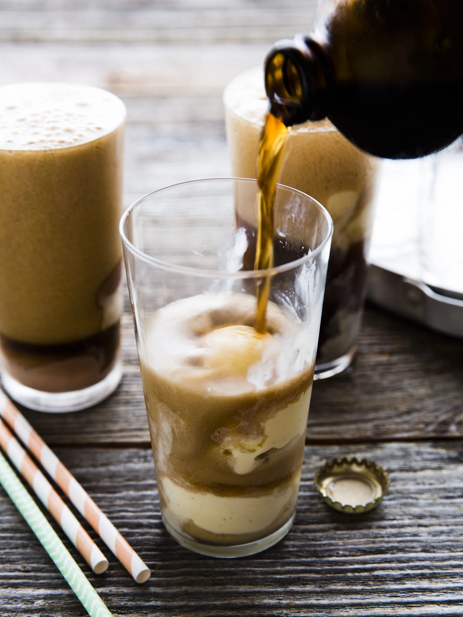 Pouring cold brew coffee into a glass over ice, with two additional glasses of iced coffee in the background on a wooden table, with colorful striped paper straws and a bottle cap nearby.