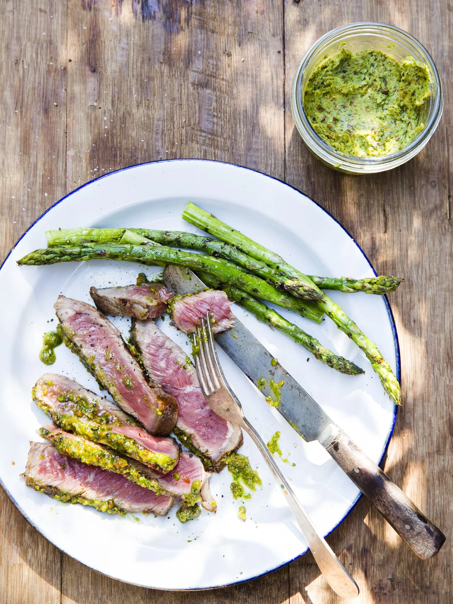 Cooked slices of steak with green sauce, grilled asparagus, and a jar of guacamole on a wooden surface.