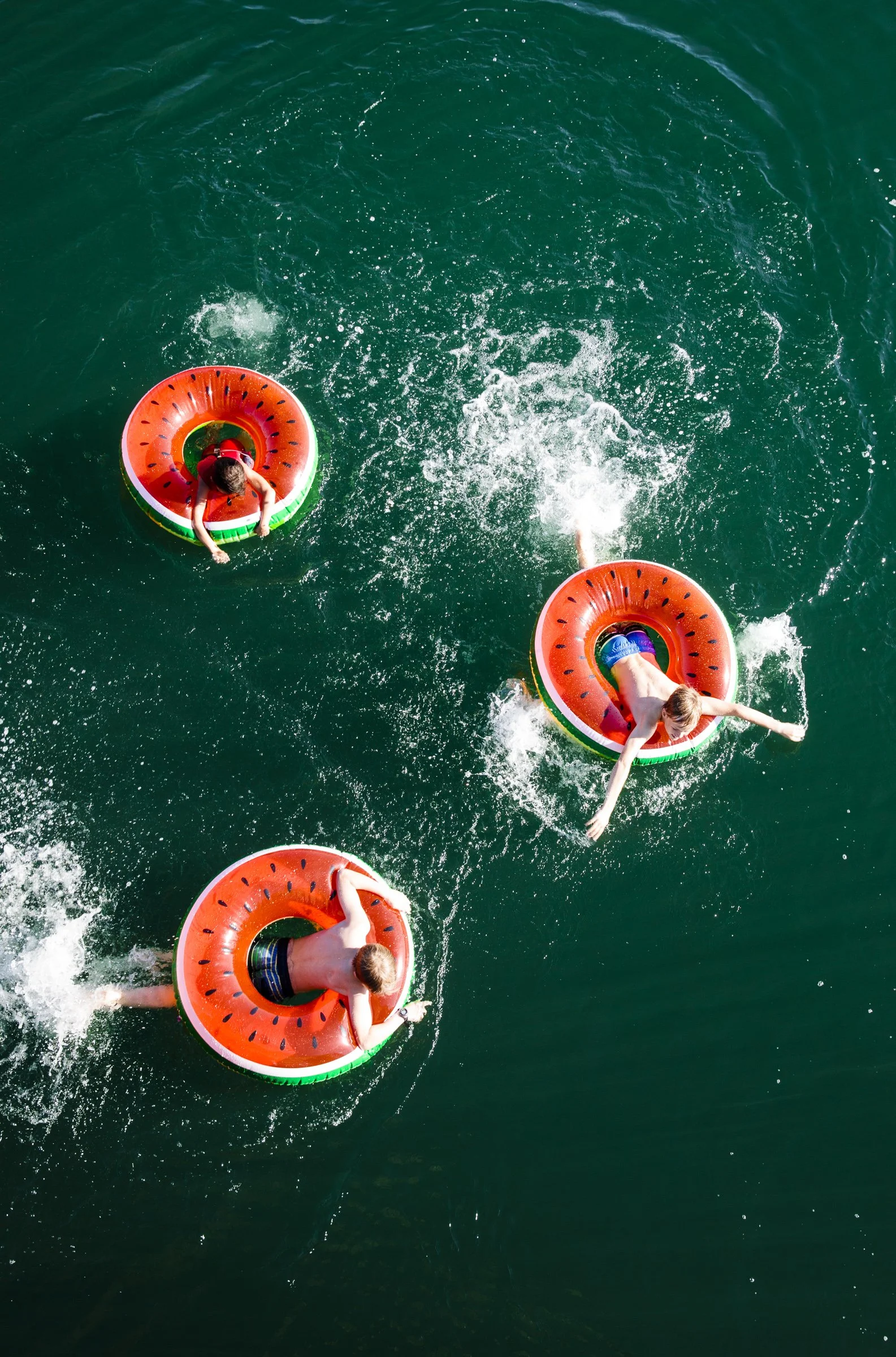 Three people floating in water on watermelon-themed inner tubes, sliding and playing.