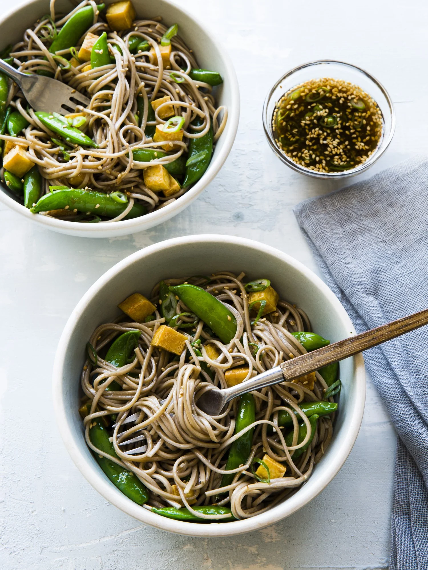Two bowls of soba noodles with snap peas and tofu with a side of dipping sauce.