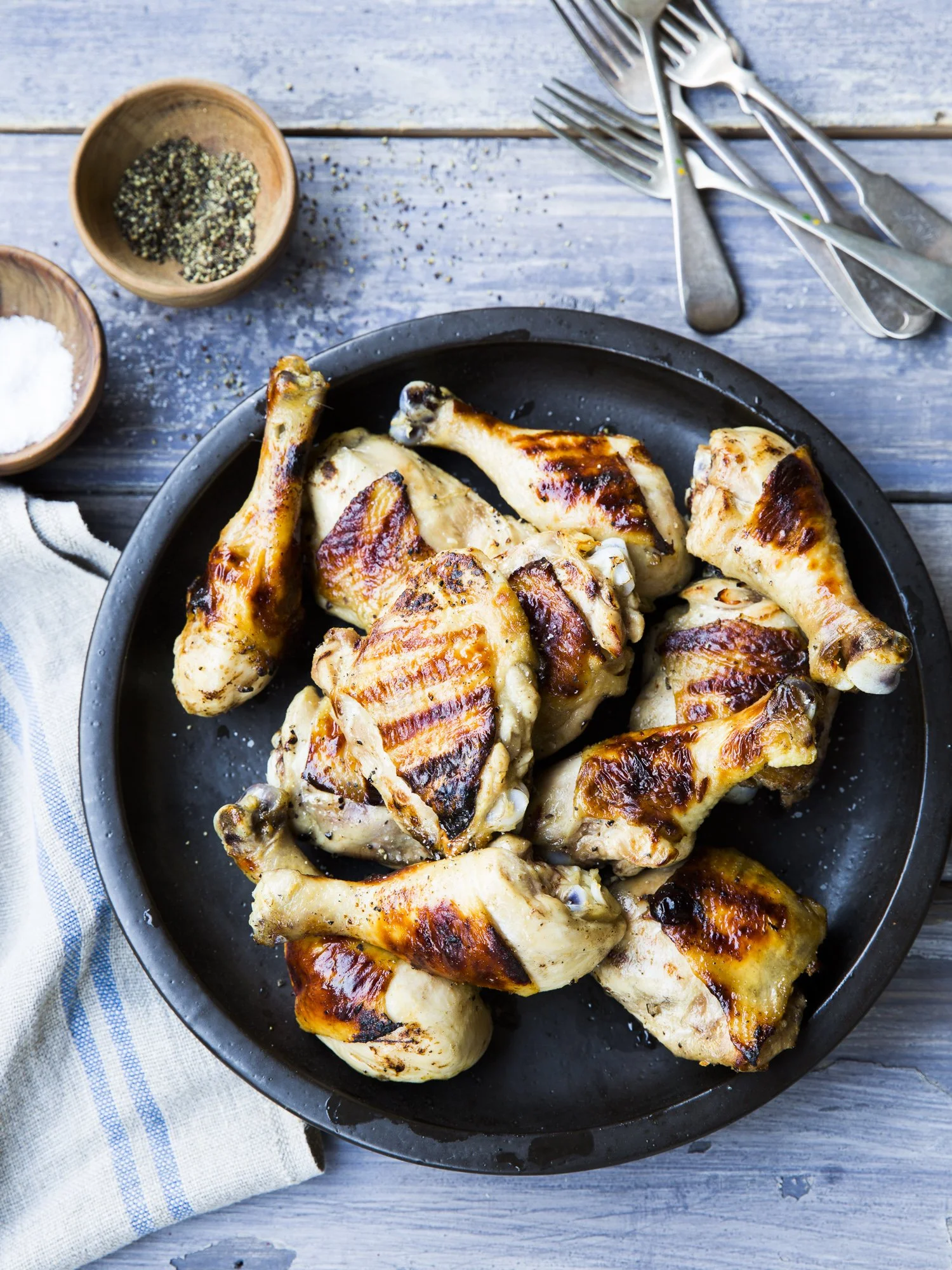 A black plate filled with grilled chicken drumsticks. The plate is on a rustic wooden table with utensils and small bowls of salt and pepper nearby.