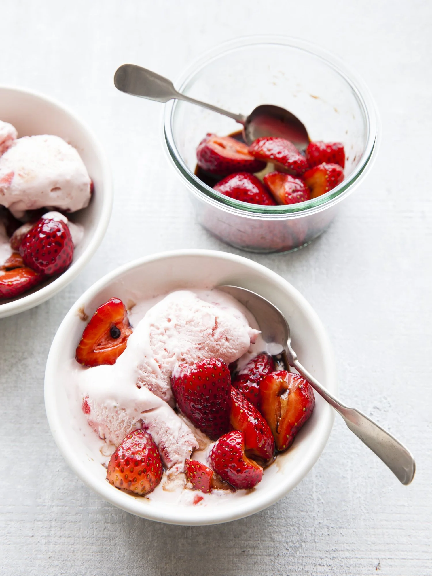 Bowl of strawberry ice cream topped with sliced strawberries, with a spoon inside, placed on a white surface. A small glass jar with chopped strawberries and a spoon, and part of another bowl of strawberry ice cream are also visible.