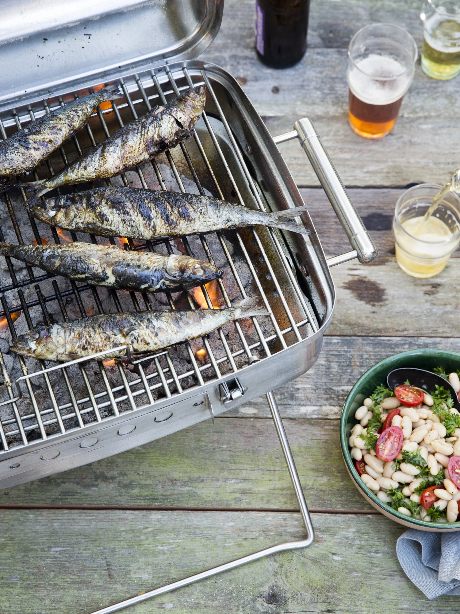 Four sardines grilling on a barbecue on a wooden table, with a bowl of bean salad, and glasses of beer and soda in the background.