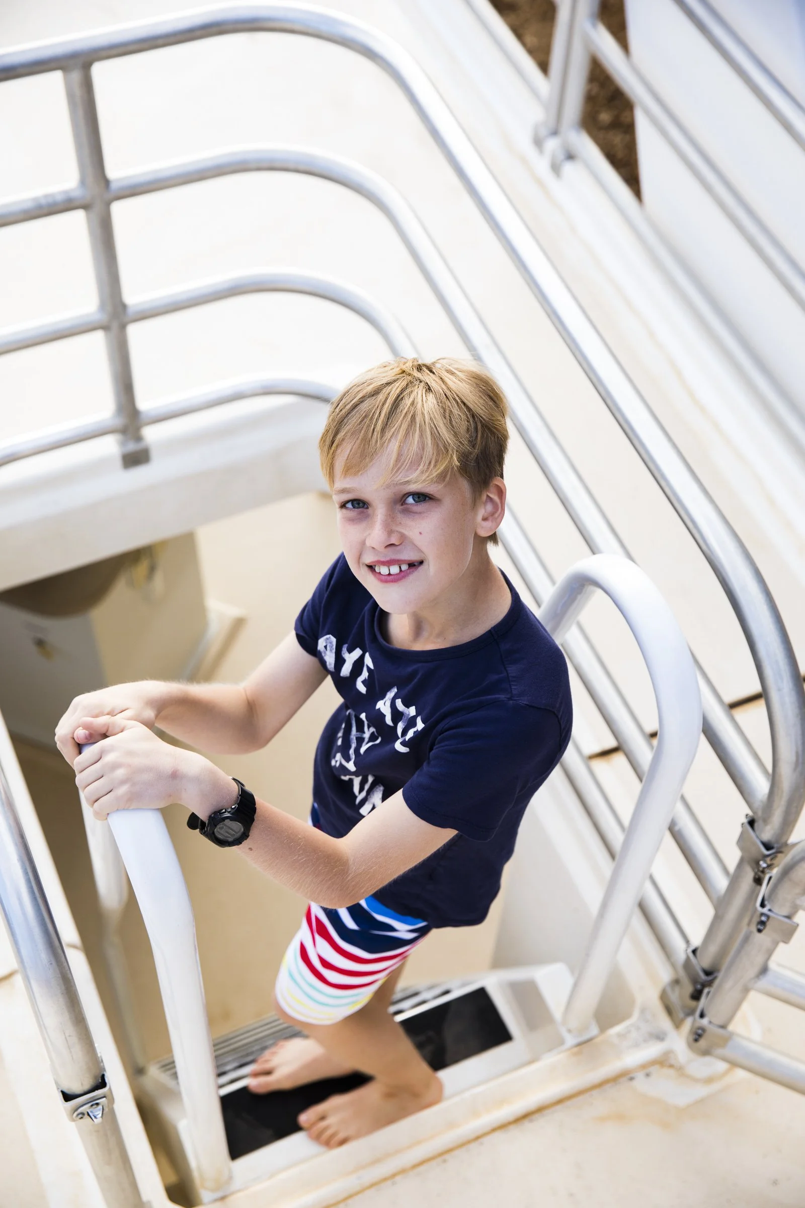 A young boy with blond hair and blue eyes standing on a staircase, looking up at the camera with a smile, holding onto the railing, wearing a navy blue t-shirt and colorful striped shorts.