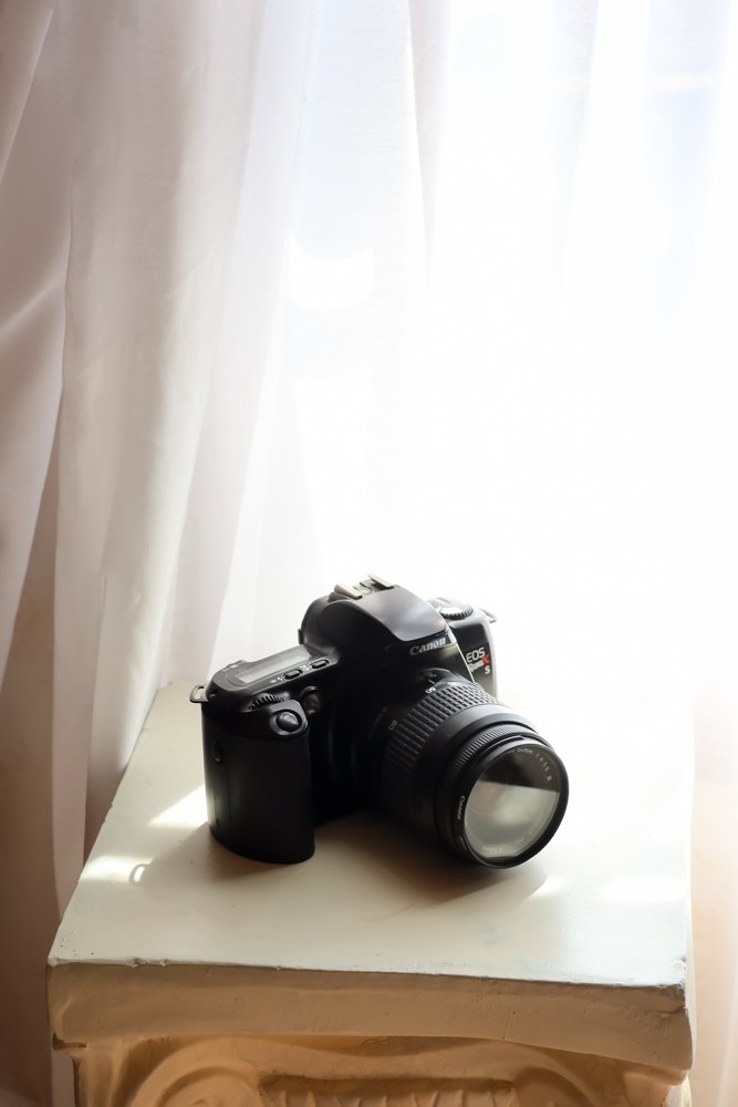 A black Canon EOS Rebel T5 camera with a lens attached, resting on a white surface near a window with cream-colored curtains.